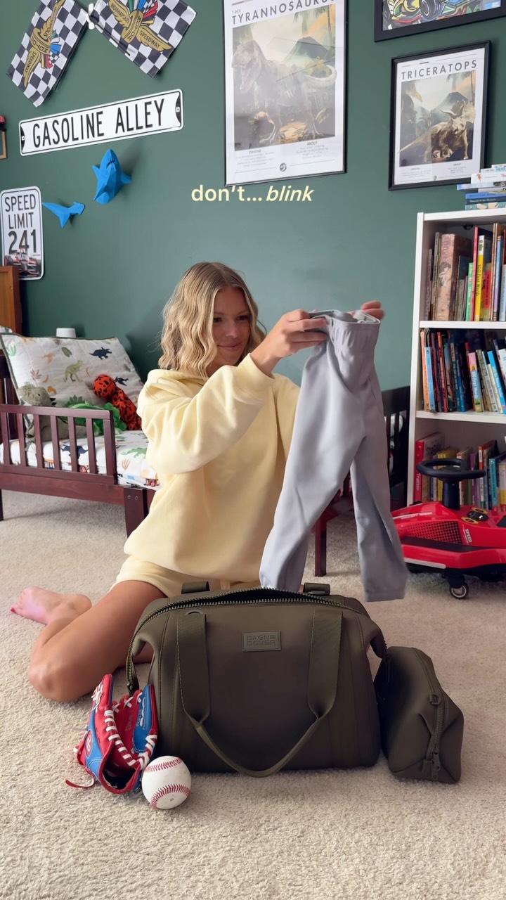 A young woman is sitting on the floor in a child's bedroom, holding up a pair of light grey pants. She is wearing a matching light yellow loungewear set. In front of her is a large olive green duffel bag, a baseball glove, and a baseball. The text "don't... blink" is overlaid on the image in white font. The background shows a child's bed with dinosaur-themed bedding, posters of dinosaurs and race cars on the wall, and a bookshelf filled with books.