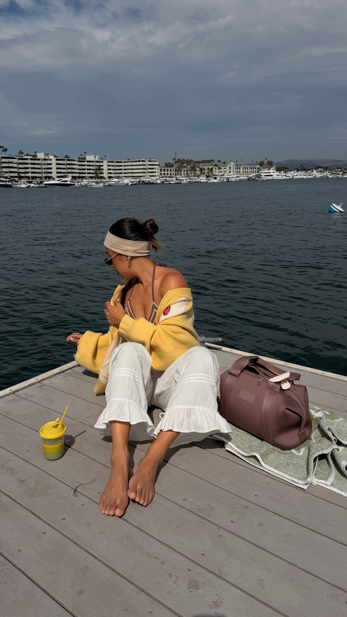 A woman sits on a wooden dock by the water, looking away from the camera. She is wearing a yellow cardigan over a striped bikini top, and white ruffled pants. Next to her is a duffel bag and a green towel. On the dock, there is also a yellow cup with a straw. The text overlay reads: "@dagnedover code: JESSBAILEY20 (20% off full priced items for 72 hours only!) We wear a lot of hats around here & the 365 Neo Collection is designed for every version of my every day. Explore the collection at @dagnedover and find the style that fits your life. #dagnedoverpartner"