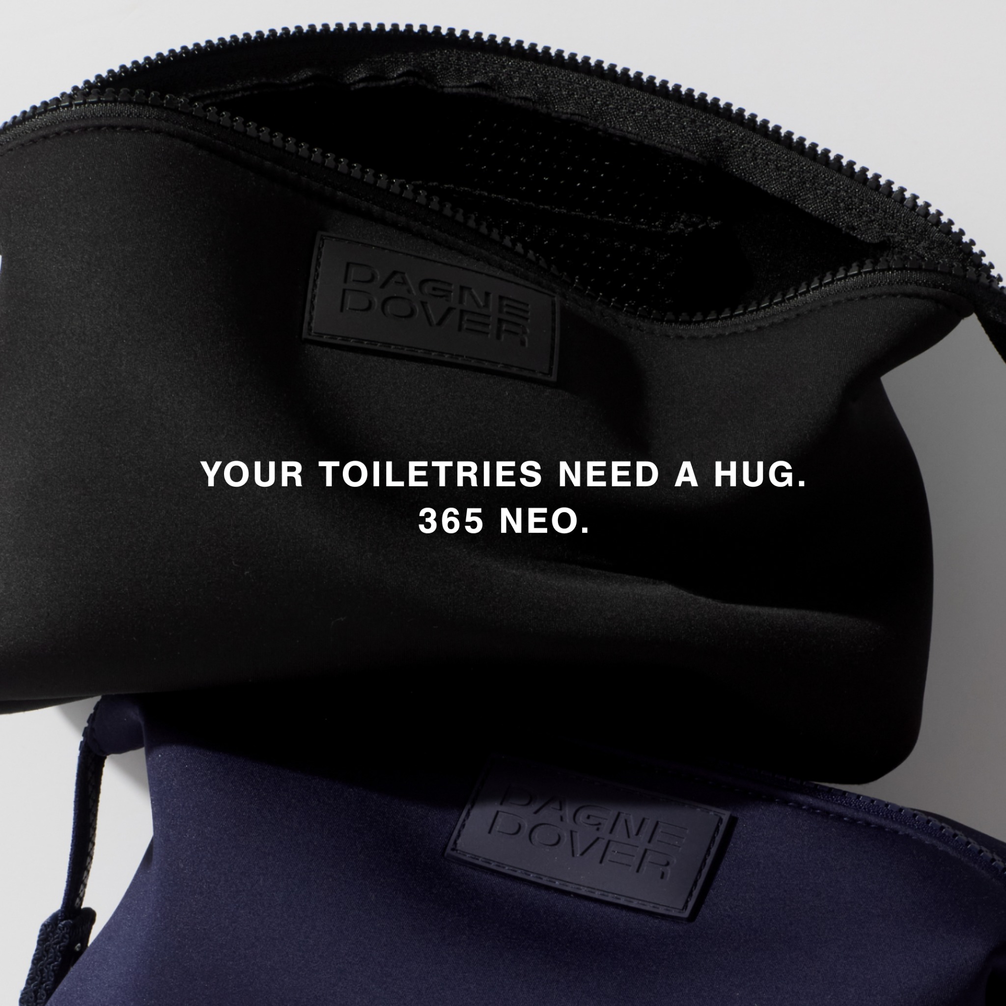 A close-up shot of two Dagne Dover toiletry bags, one black and one navy blue, are stacked on top of each other. The black bag is slightly open, revealing its interior. White text is overlaid on the image, with the main headline reading "YOUR TOILETRIES NEED A HUG." Below this, in smaller text, it says "365 NEO." The overall aesthetic is clean and minimalist, focusing on the product and the message.