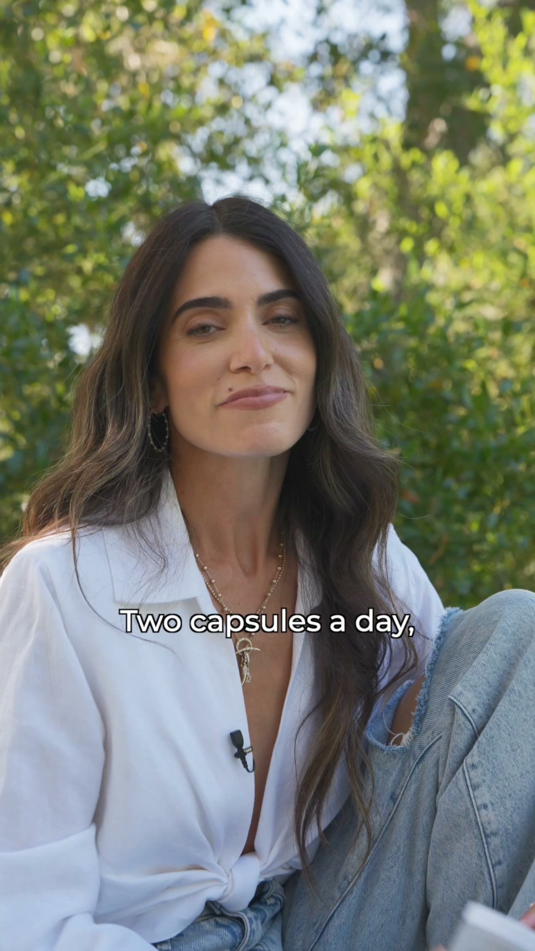 A woman with long, wavy brown hair is sitting outdoors, wearing a white button-down shirt and ripped jeans. She is looking towards the camera with a slight smile. Text overlay on the image reads "Two capsules a day,". The background is blurred greenery, suggesting a natural setting. The overall impression is a lifestyle shot with an informational overlay.