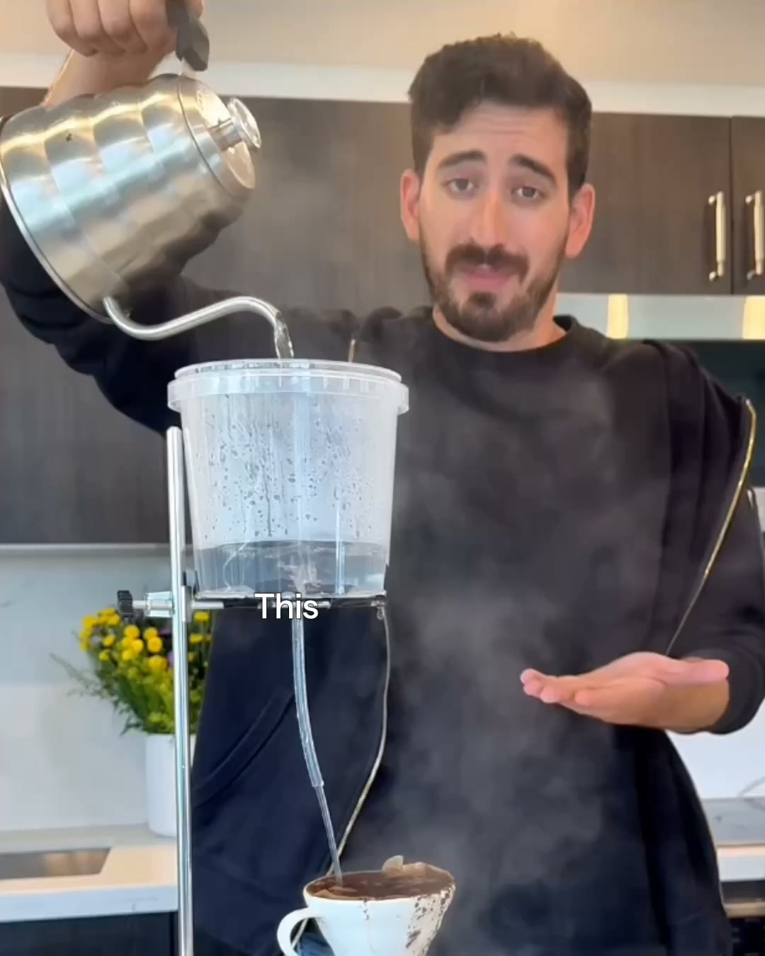A man in a black hoodie is pouring hot water from a stainless steel kettle into a clear plastic container with a metal stand. The container has a tube leading down to a coffee filter and a white mug, where coffee is brewing. Steam rises from the setup. The text "This" is overlaid on the plastic container. The man looks at the camera with a slight smile and his hand outstretched. The background shows a kitchen counter and cabinets.