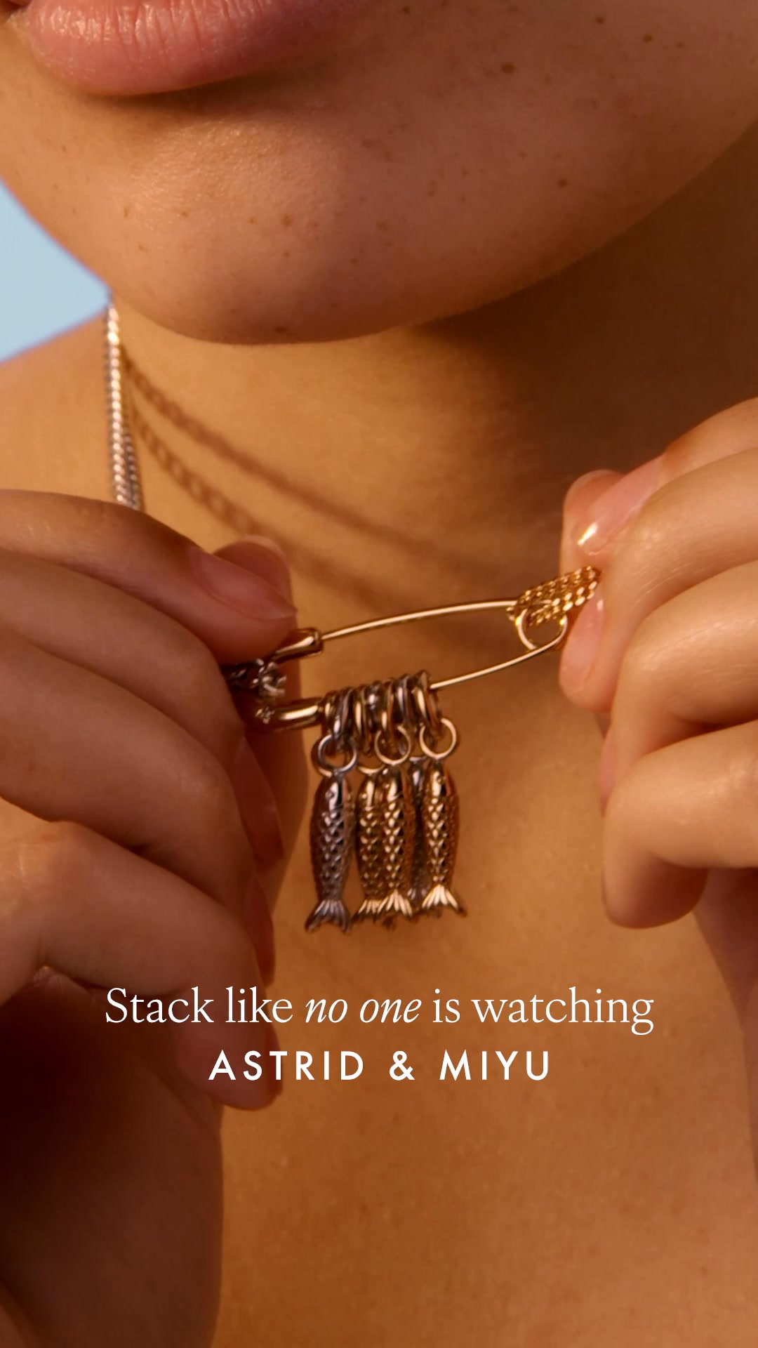 A close-up shot focuses on a person's neck and collarbone, where they are holding a gold safety pin with three small, dangling fish charms. The charms are silver and gold, with intricate scale details. The text "Stack like no one is watching" is overlaid in white, followed by the brand name "ASTRID & MIYU" in a slightly larger, bolder font. The background is a soft, out-of-focus blue and warm skin tones.