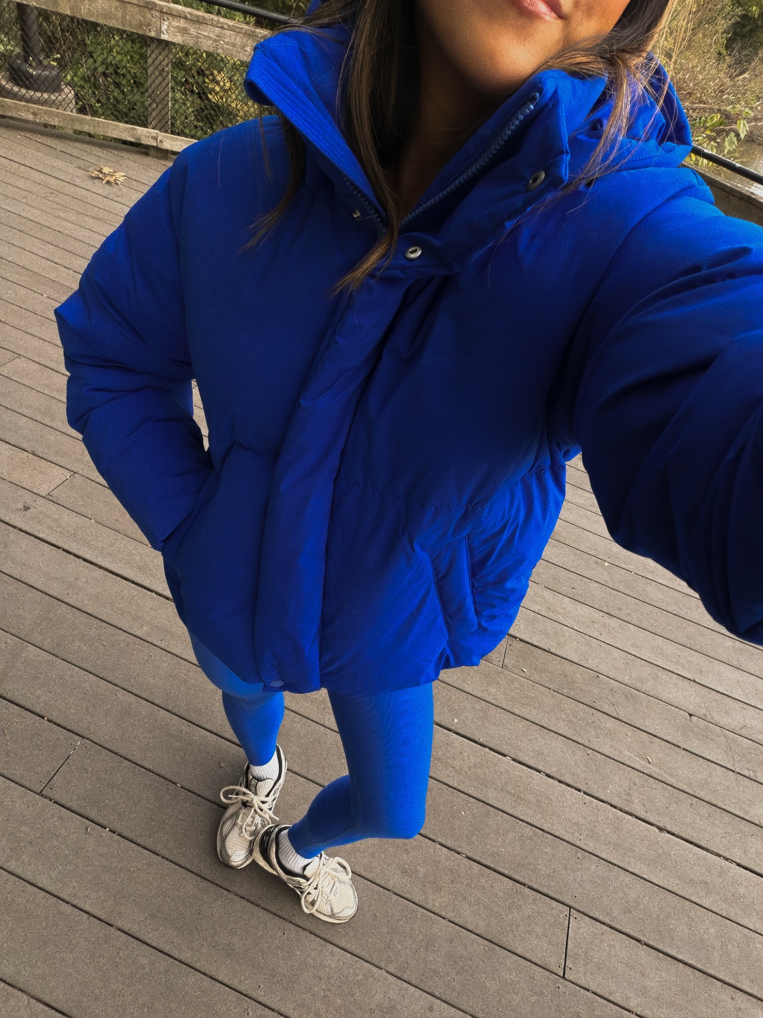 A woman is shown from the chest down, wearing a bright blue puffer jacket and matching blue leggings. She is standing on a wooden boardwalk. The text overlay reads "She’s not like a regular blue ... she’s Ultra Blue. 💙 vibes via @jenniferrsweet". The image is a selfie taken from a high angle, focusing on the outfit.