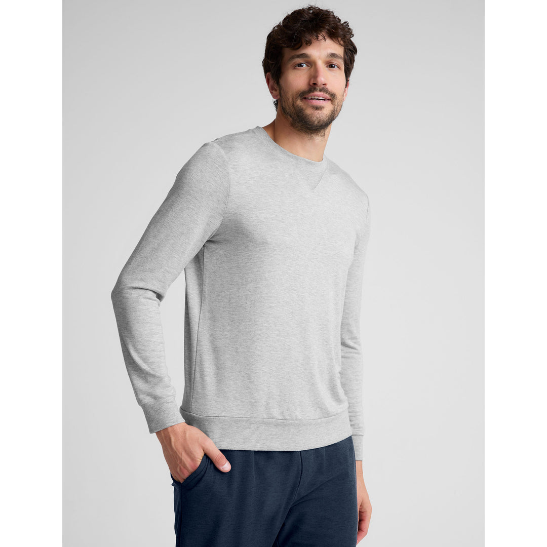 A man with short brown hair and a beard, wearing a light grey long-sleeved crewneck shirt and dark blue sweatpants, stands against a plain grey background. He is looking towards the camera with a slight smile, his left hand in his pocket. White text overlaid on the image reads "Reimagined essentials designed to perform. Shop our Men's Collection today."