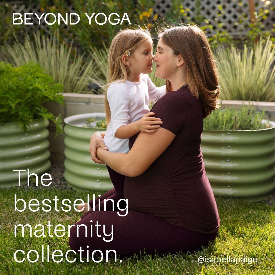 A pregnant woman kneels on grass, holding a young child. The woman is wearing a dark-colored maternity outfit, and the child is wearing a white long-sleeved shirt. They are nose-to-nose, sharing an intimate moment. White text overlays the image, reading "BEYOND YOGA" at the top and "The bestselling maternity collection." at the bottom left. The Instagram handle "@isabellapaige_" is visible in the bottom right corner.