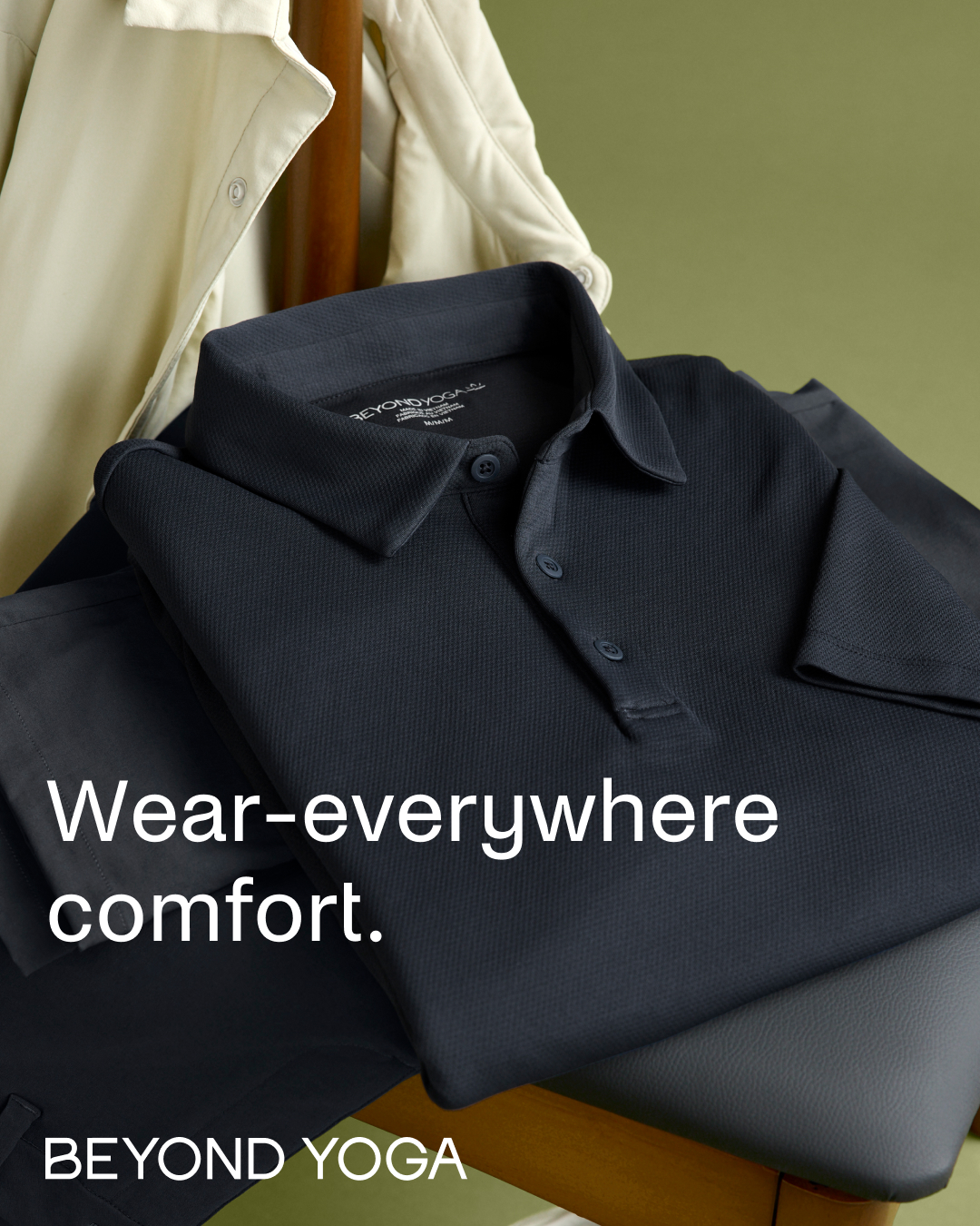 A dark charcoal polo shirt is folded neatly on top of dark pants, with a cream-colored trench coat draped in the background. The text "Wear-everywhere comfort." is overlaid in large white sans-serif font across the bottom half of the image. Below this, the brand name "BEYOND YOGA" is displayed in white capital letters. The overall aesthetic is clean and focuses on the comfort and versatility of the apparel.