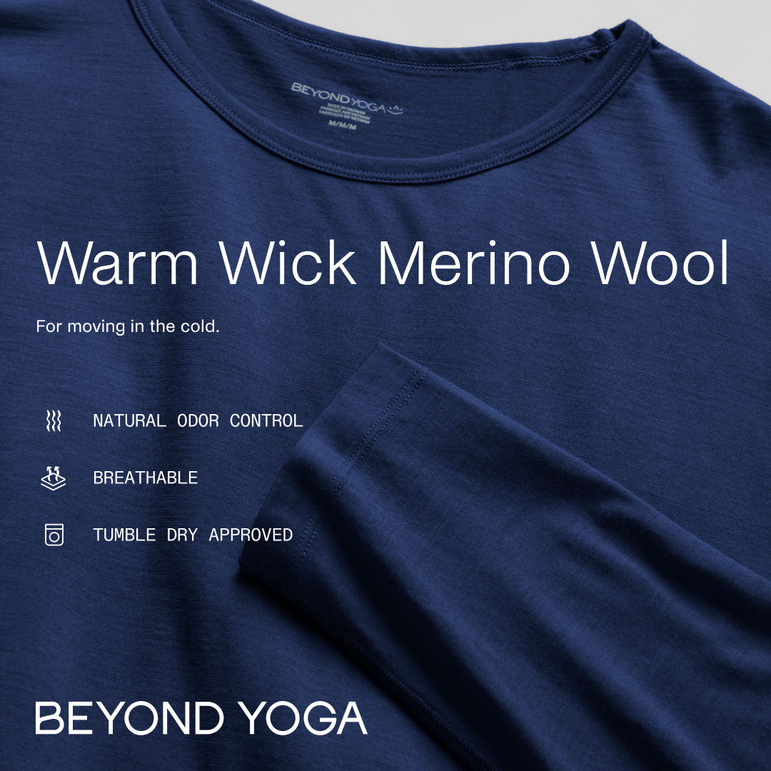 A close-up shot of a navy blue long-sleeved shirt from Beyond Yoga. The text "Warm Wick Merino Wool" is overlaid in large white font. Below that, in smaller white font, it reads "For moving in the cold." To the left, three icons with text explain the benefits: "NATURAL ODOR CONTROL," "BREATHABLE," and "TUMBLE DRY APPROVED." The brand name "BEYOND YOGA" is displayed in large white letters at the bottom of the image.