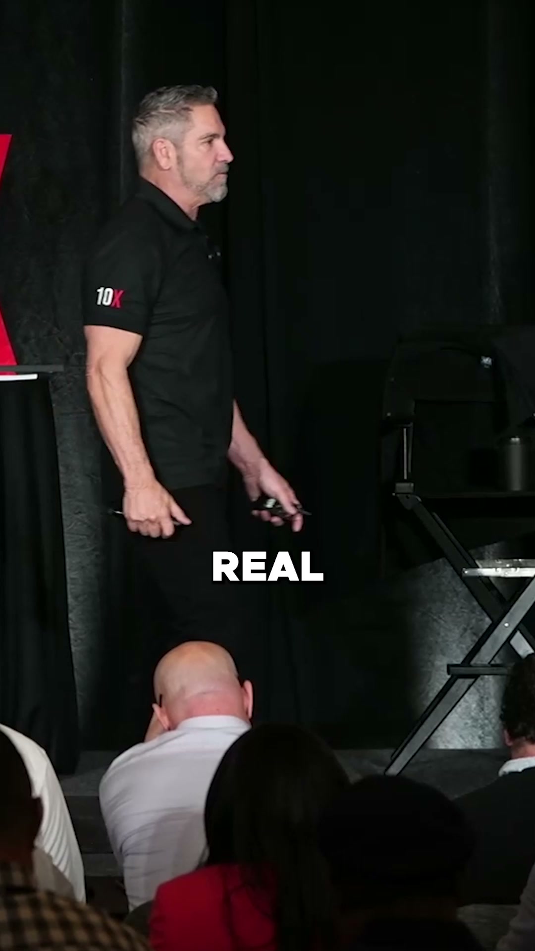 A man in a black polo shirt with a "10X" logo on the sleeve stands on a stage, speaking to an audience. He is holding a microphone in his right hand and a small device in his left. The word "REAL" is displayed in large white capital letters across the lower-middle section of the image. The audience members are seated in the foreground, mostly with their backs to the camera. The background is dark and appears to be a stage curtain.