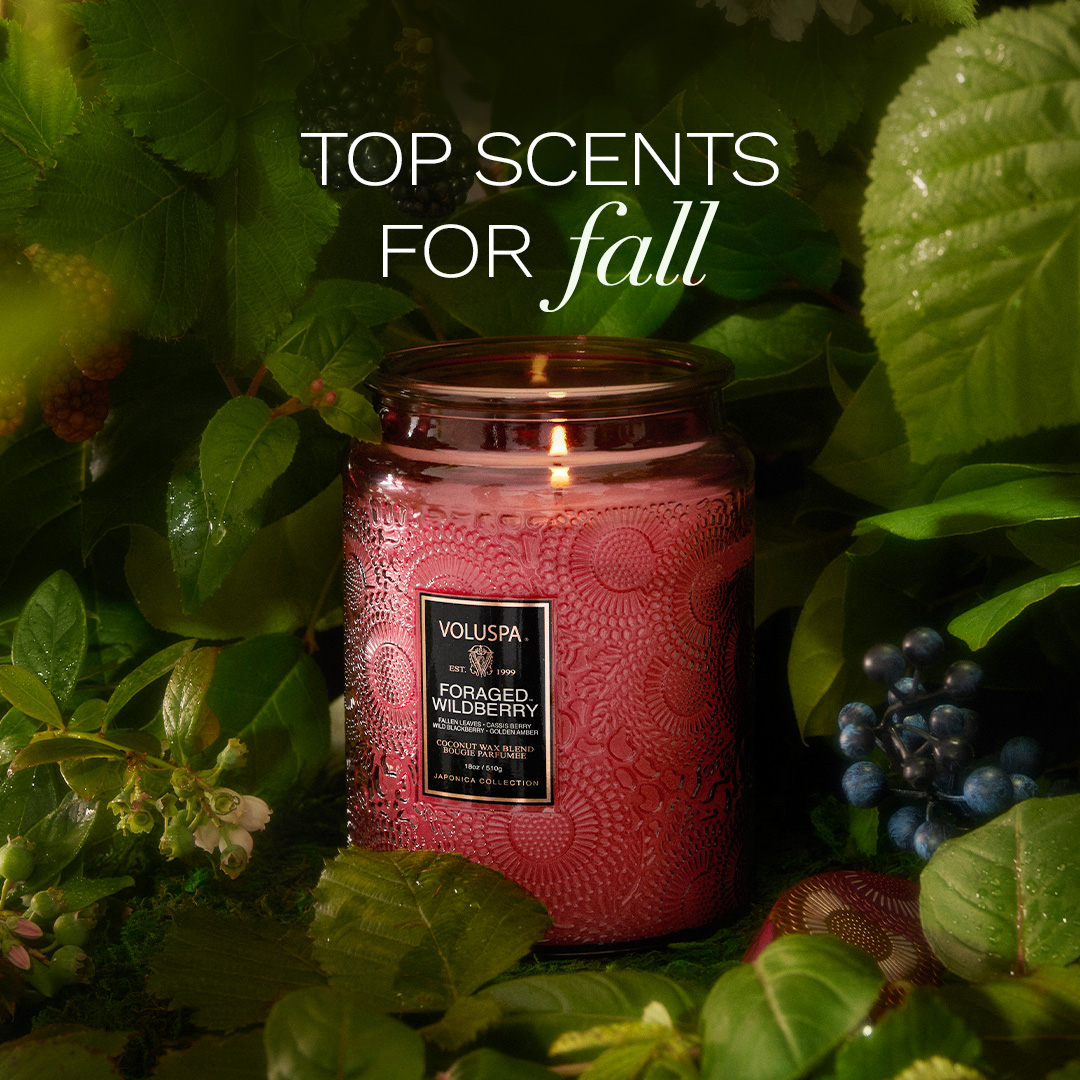 A lit Voluspa candle with a red, embossed jar sits in the center of the frame, surrounded by lush green leaves and dark berries. The text "TOP SCENTS FOR fall" is overlaid at the top of the image in white and script font. The candle label reads "VOLUSPA FORAGED. WILDBERRY". The overall aesthetic evokes a cozy and autumnal atmosphere, highlighting the candle as a perfect scent for the fall season.