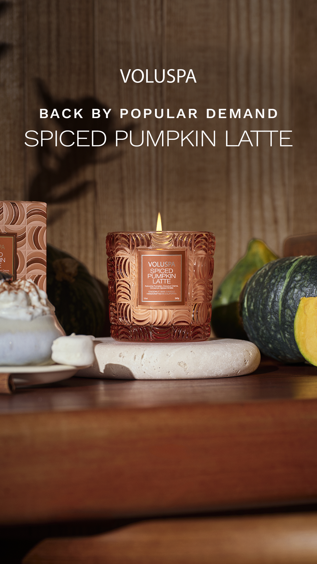 A lit Voluspa Spiced Pumpkin Latte candle is displayed prominently on a wooden surface, surrounded by decorative pumpkins and a whipped cream dessert. The candle jar is ornate and amber-colored, with the label clearly visible. Text overlays the image, announcing "VOLUSPA" at the top, followed by "BACK BY POPULAR DEMAND" and "SPICED PUMPKIN LATTE" in large, white, sans-serif font. The overall aesthetic evokes a cozy, autumnal atmosphere.