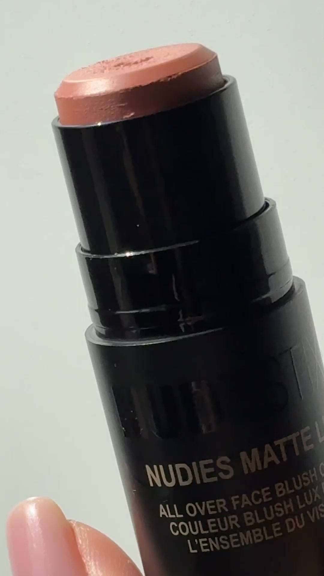 A close-up shot of a Nudestix Nudies Matte Lux stick in the shade Nude Buff. The product is black with silver text that reads "NUDIES MATTE LUX ALL OVER FACE BLUSH LUX". The hook text "If you love a no-makeup look, powder blush can make skin look heavy." is overlaid on the image. The text explains how powder blush can look heavy and settle into texture, contrasting it with the desired soft, bare look.