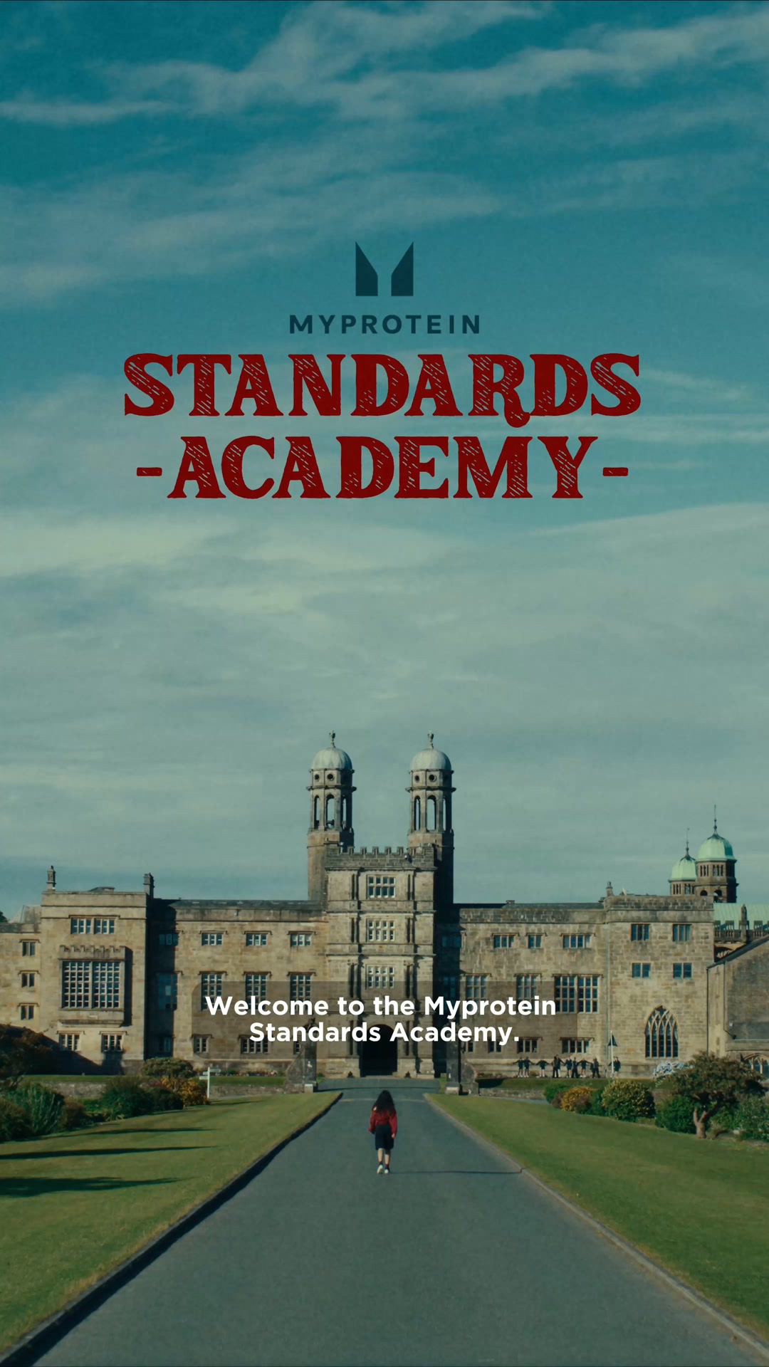 A wide shot shows a grand, old building with towers and many windows, set against a cloudy blue sky. A long, paved driveway leads up to the building, with a lone figure walking away from the camera towards it. The text "MYPROTEIN STANDARDS -ACADEMY-" is displayed prominently in large, red, distressed lettering at the top of the image. Below the building, white text reads "Welcome to the Myprotein Standards Academy."