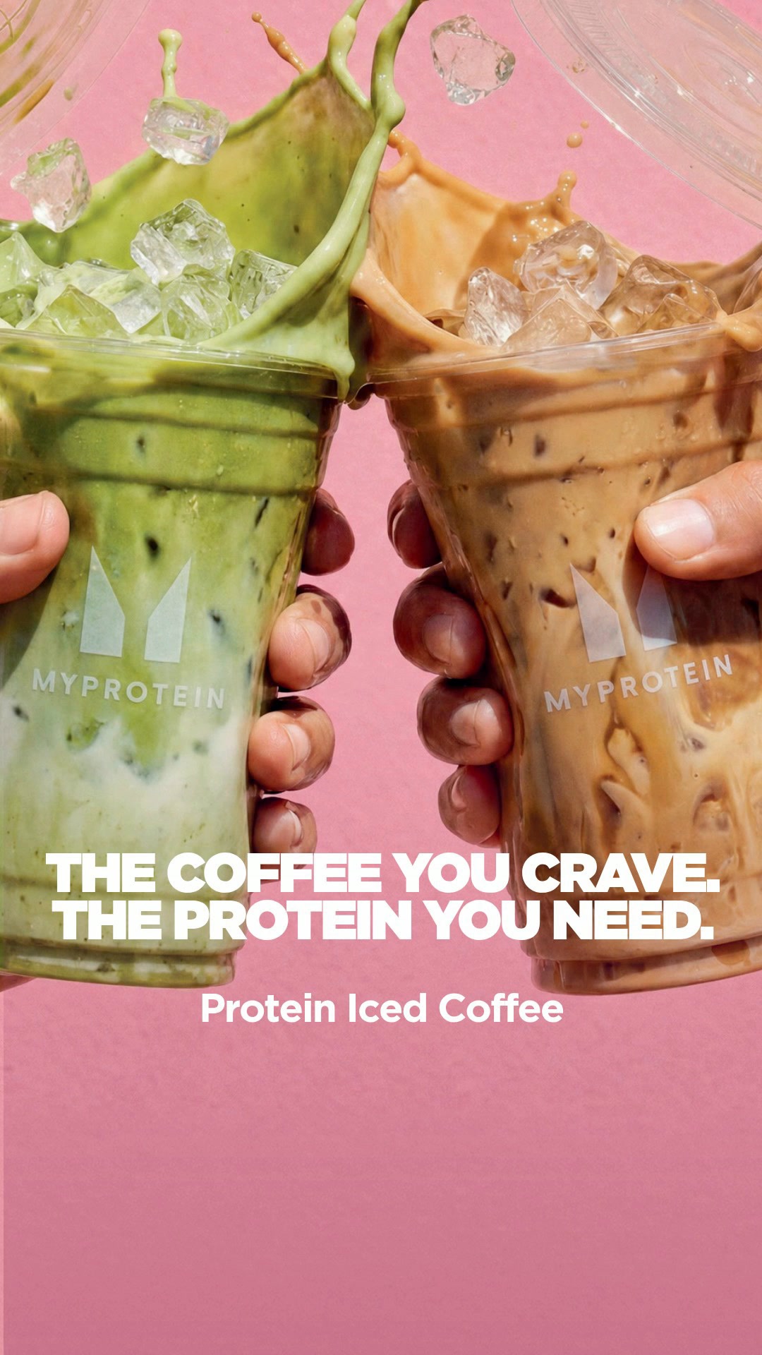 Two hands hold up clear plastic cups filled with iced coffee drinks, one a creamy beige color and the other a vibrant green, with ice cubes splashing out of both. The MyProtein logo is visible on both cups. White text overlays the bottom half of the image, reading "THE COFFEE YOU CRAVE. THE PROTEIN YOU NEED." followed by "Protein Iced Coffee" on a second line. The background is a solid, bright pink color.