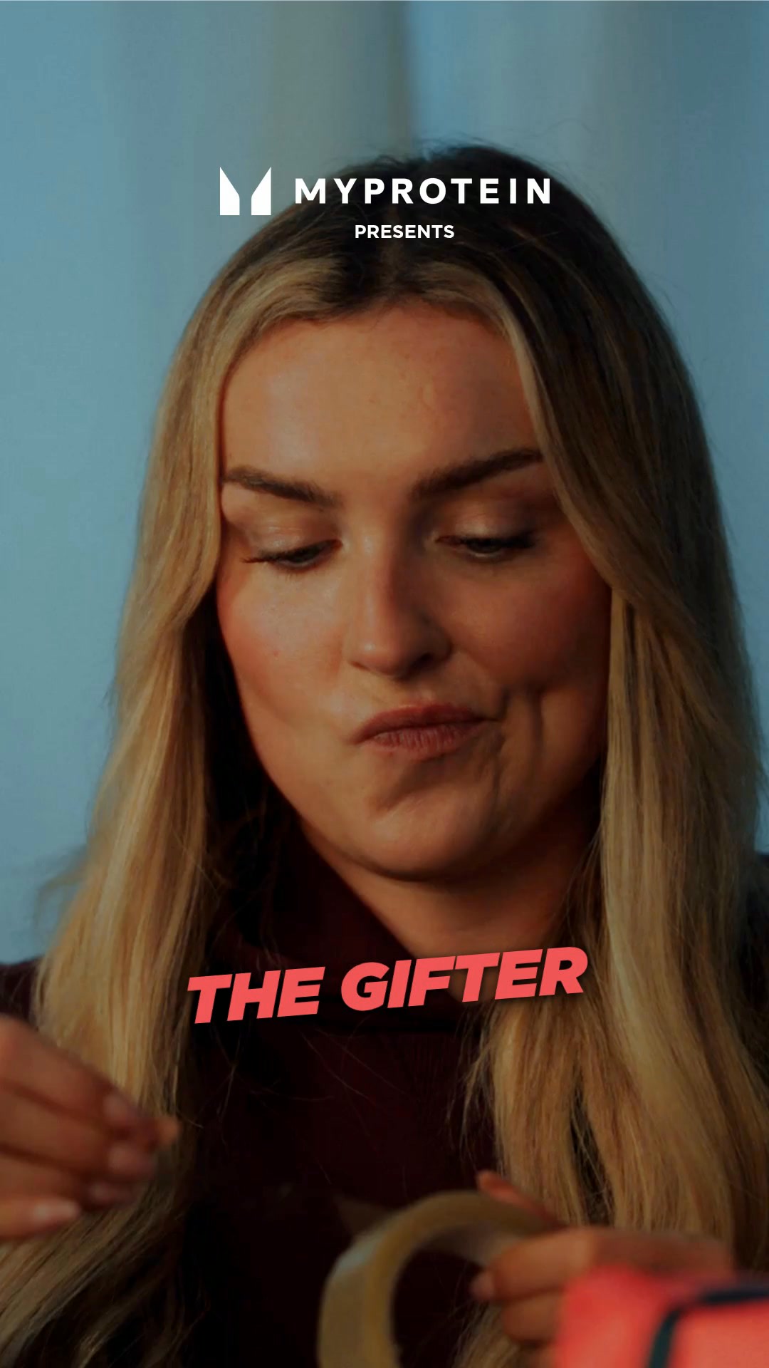 A close-up shot of a blonde woman with her hair down, looking down and to the side with a slightly pursed-lip expression. She is in the process of wrapping a gift, holding a roll of tape. Large, bold, pink text overlay reads "THE GIFTER" across the lower portion of the image. The MyProtein logo and "PRESENTS" are visible in the upper left corner. The ad context suggests this is related to Black Friday deals.