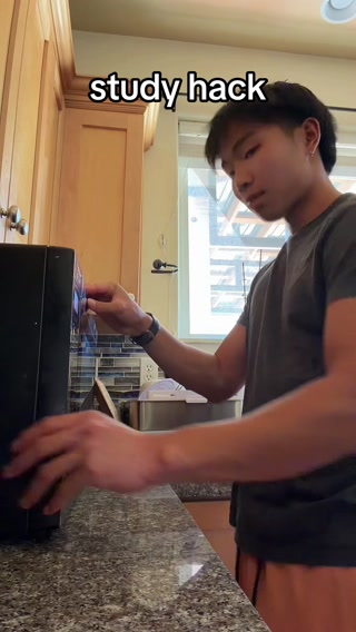 A young Asian man is shown in a kitchen, reaching towards a microwave. Text overlayed on the top of the image reads "study hack". The man is wearing a dark t-shirt and orange shorts. The kitchen has wooden cabinets and a granite countertop. The overall scene suggests a casual, everyday setting where a productivity tip is being shared.