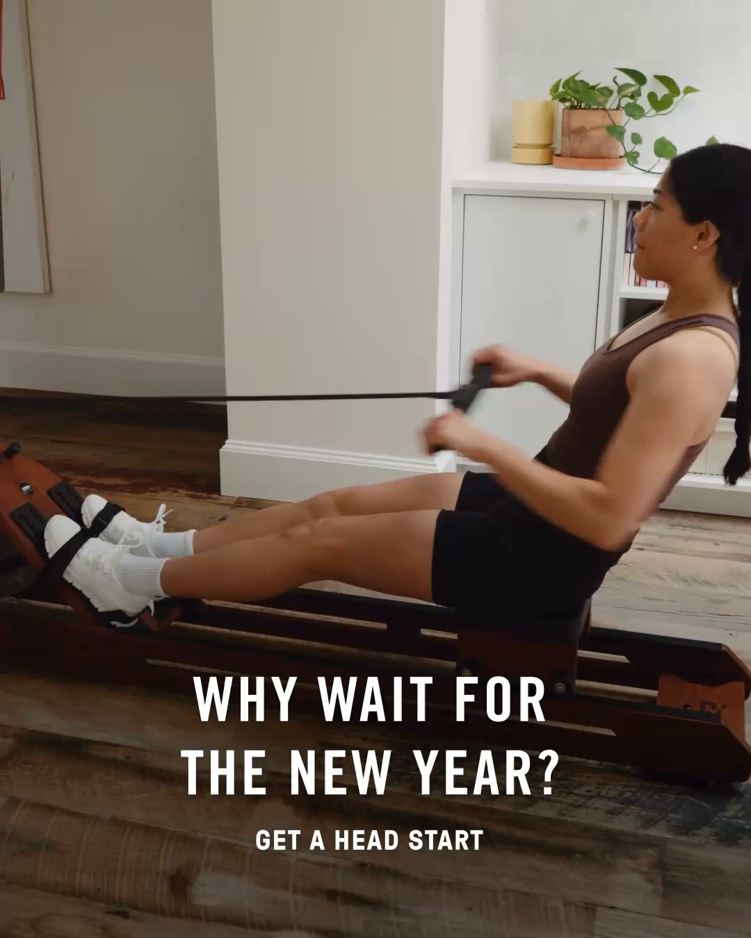 A woman is shown from the side, rowing on a rowing machine in a home setting. The text on the screen asks, "WHY WAIT FOR THE NEW YEAR?" Below this, in smaller text, it says, "GET A HEAD START." The overall scene suggests fitness and starting a new routine before the traditional New Year's resolution period.