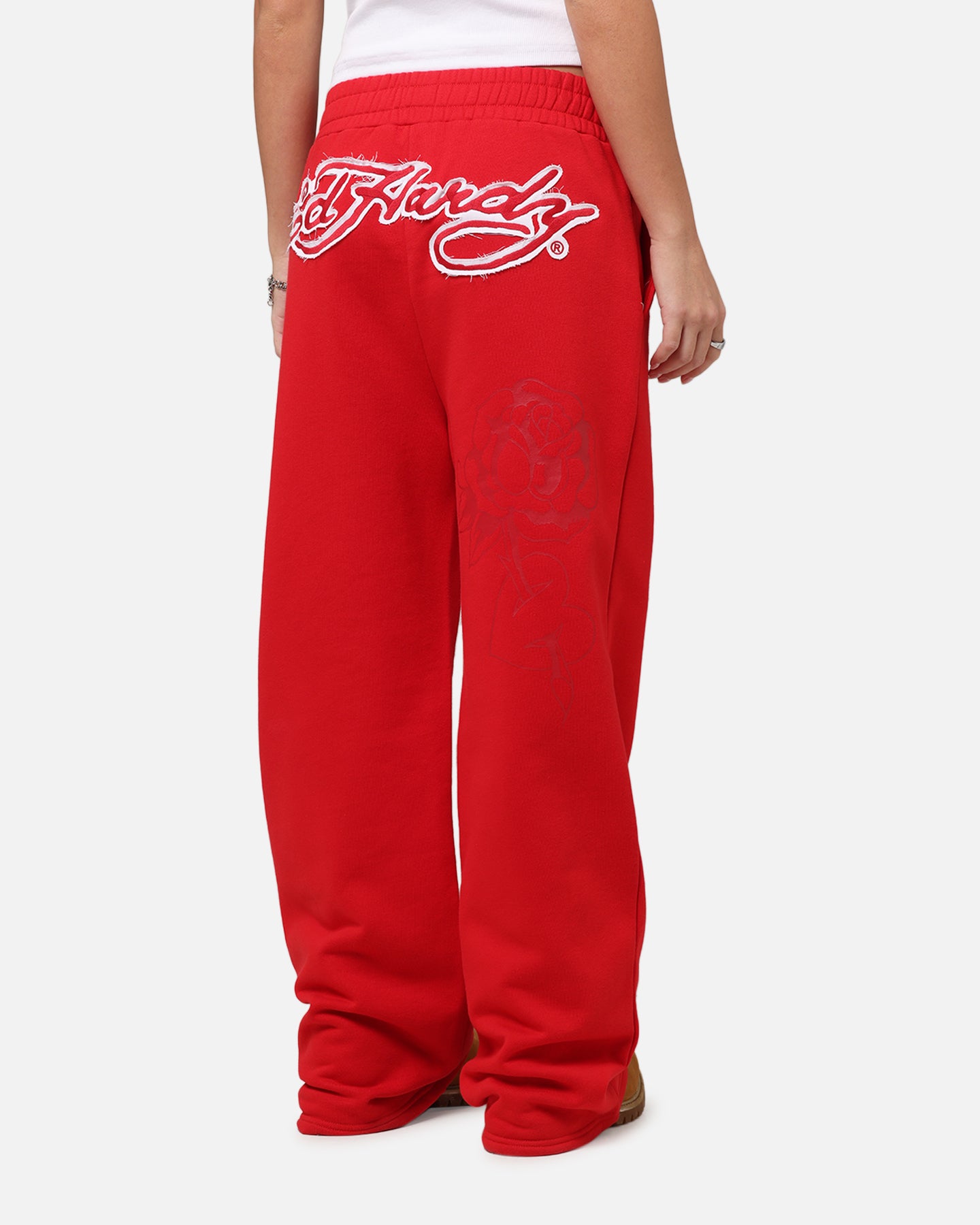 A person is shown from the back, wearing bright red sweatpants with a large white Ed Hardy logo emblazoned across the upper back. A subtle, tonal rose graphic is visible on the left leg of the sweatpants. The text "Your next fit is waiting" is overlaid on the image, accompanied by an emoji.