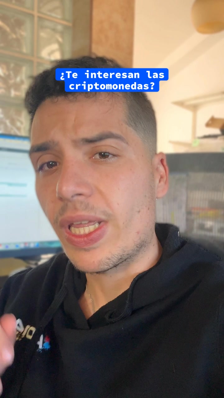 A close-up shot of a man's face with a black hoodie. He is looking directly at the camera and speaking. Blue text overlay at the top of the screen asks, "¿Te interesan las criptomonedas?" which translates to "Are you interested in cryptocurrencies?". The background is blurred, showing what appears to be an office or home environment with a computer monitor and shelves.