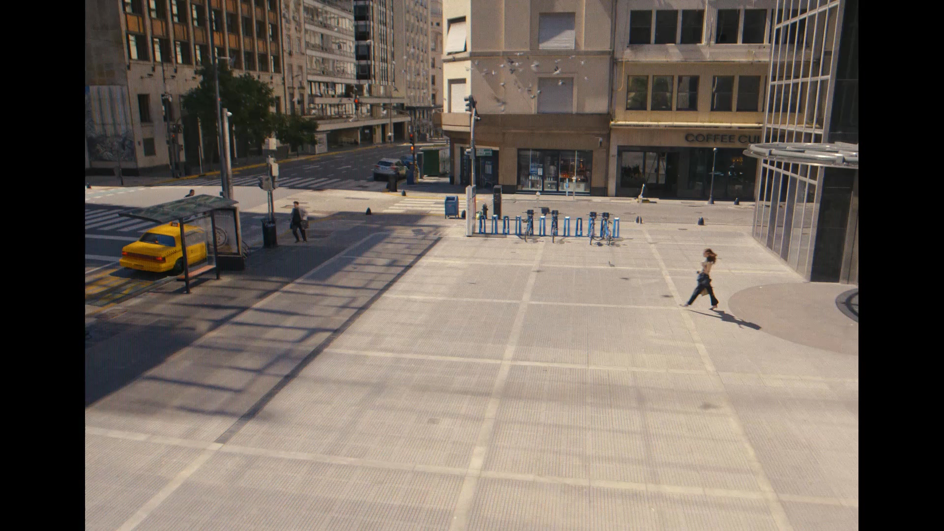 An aerial view of a city square shows a yellow taxi at a bus stop on the left. A person walks across the square on the right. In the background, buildings line the street, and a coffee shop with the words "COFFEE CU" is visible. The text "Ready to take the first step out of the broken financial system?" is overlaid on the image.