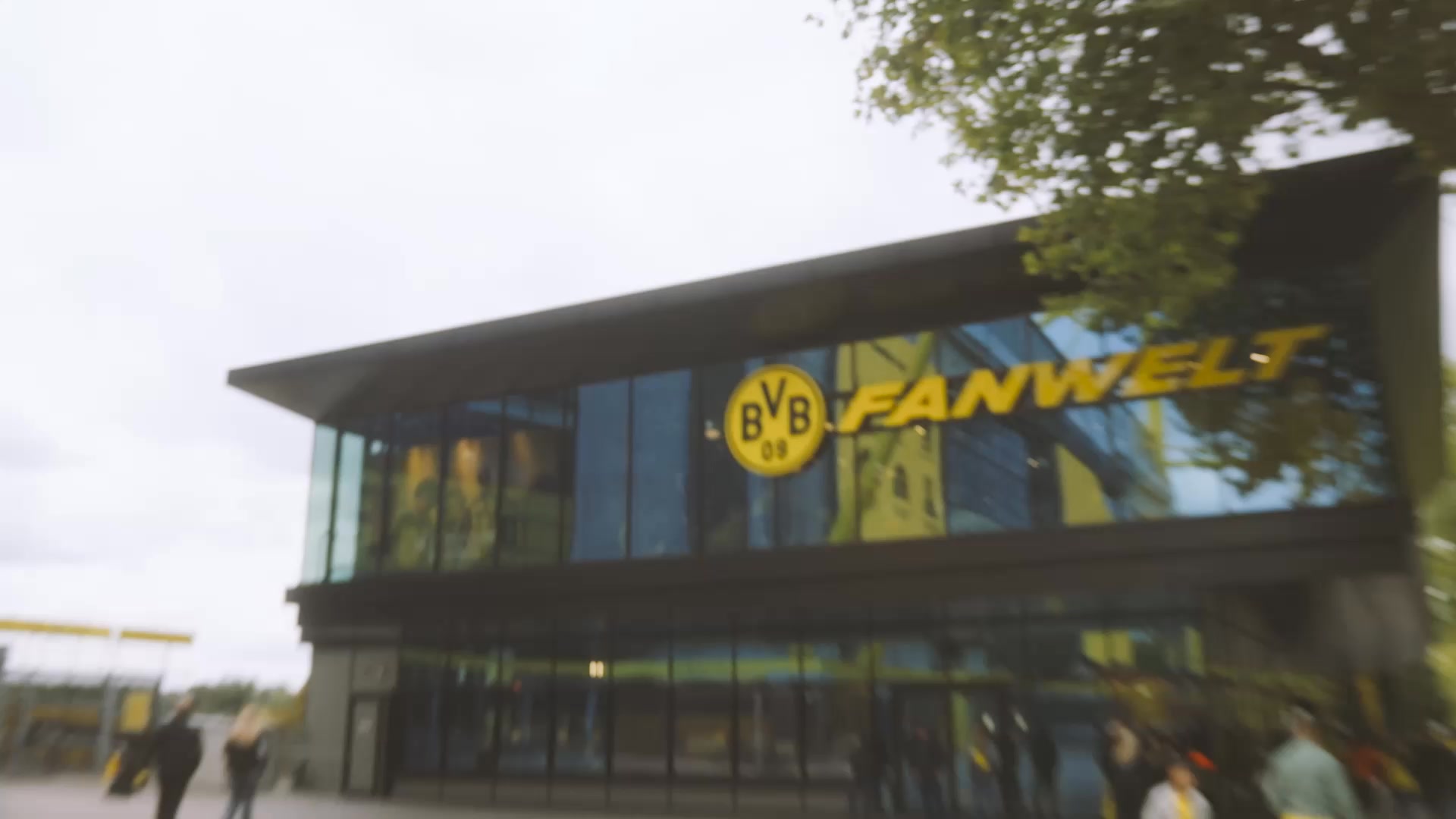 The creative shows the exterior of a modern building with large glass windows. The building has a dark facade and a prominent yellow logo that reads "BVB FANWELT". The text "Krypto kaufen, verkaufen und verwenden mit Coinbase – der sicheren App für Ihr Krypto." is overlaid on the image. The overall impression is of a commercial establishment, possibly a fan store or a club headquarters, with the ad copy promoting a cryptocurrency service.