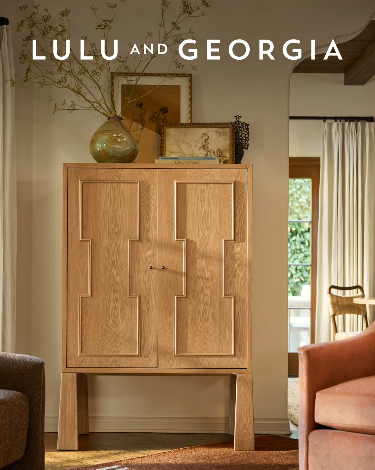 The creative is a lifestyle image with text overlay. It features a beautifully crafted wooden cabinet with intricate geometric carvings on its doors. The cabinet is placed in a well-decorated room, with a vase of dried flowers on top and artwork displayed behind it. The text "LULU AND GEORGIA" is prominently displayed at the top of the image. The hook "Hidden in plain sight, ready when you are." is positioned below the brand name, creating a sense of intrigue and anticipation about the product.