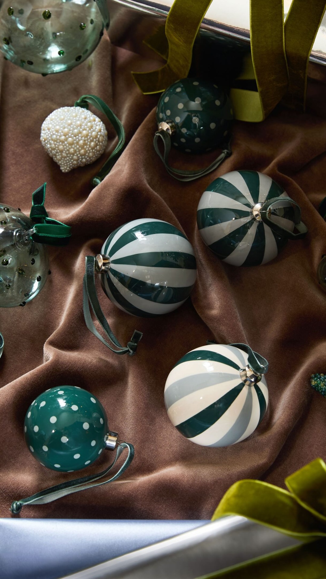 A flatlay arrangement of various Christmas ornaments on a plush, brown velvet fabric. The ornaments include striped baubles in green and white, a green bauble with white polka dots, and a white bauble covered in small pearls. Green velvet ribbons are draped around the ornaments, adding a festive touch. The text "You could say it’s one of our best ornament curations yet." is overlaid on the image.