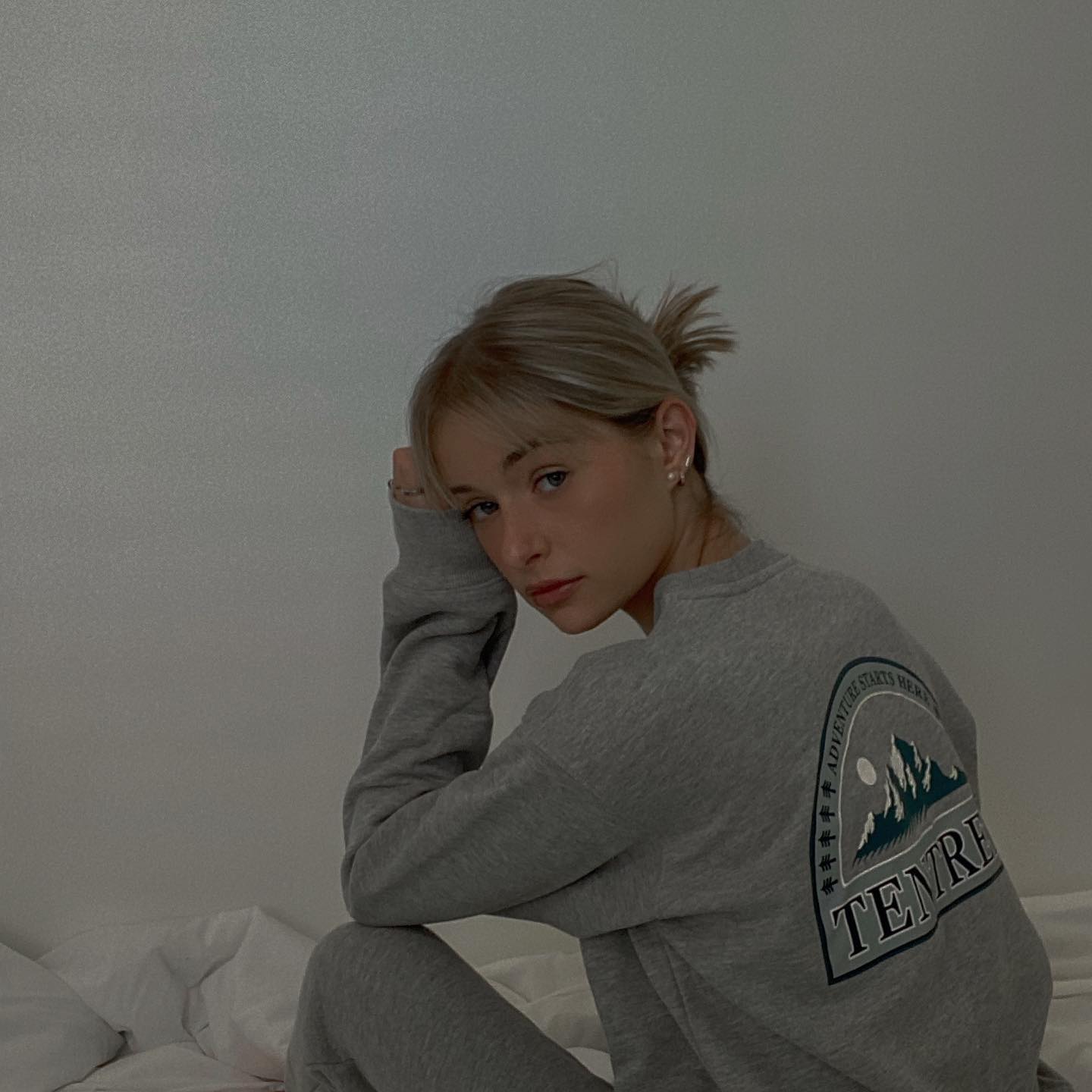 A young woman with blonde hair pulled back in a messy bun is sitting on a bed, leaning her head on her hand. She is wearing a grey sweatshirt with a graphic design on the back that says "ADVENTURE STARTS HERE" and features mountains. The text "TENRE" is also visible on the graphic. The background is a plain, light-colored wall. The hook text "the coziest set from @tentree" is overlaid on the image.