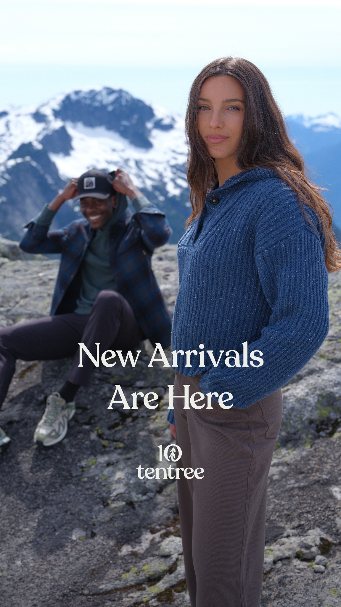 A woman in a blue sweater and brown pants stands in the foreground, looking at the camera. Behind her, a man in a plaid shirt and hoodie sits on rocks, adjusting his baseball cap. They are in a mountainous landscape with snow-capped peaks in the background. White text overlaid on the image reads "New Arrivals Are Here" and the Tentree logo with the text "10 tentree" below it.