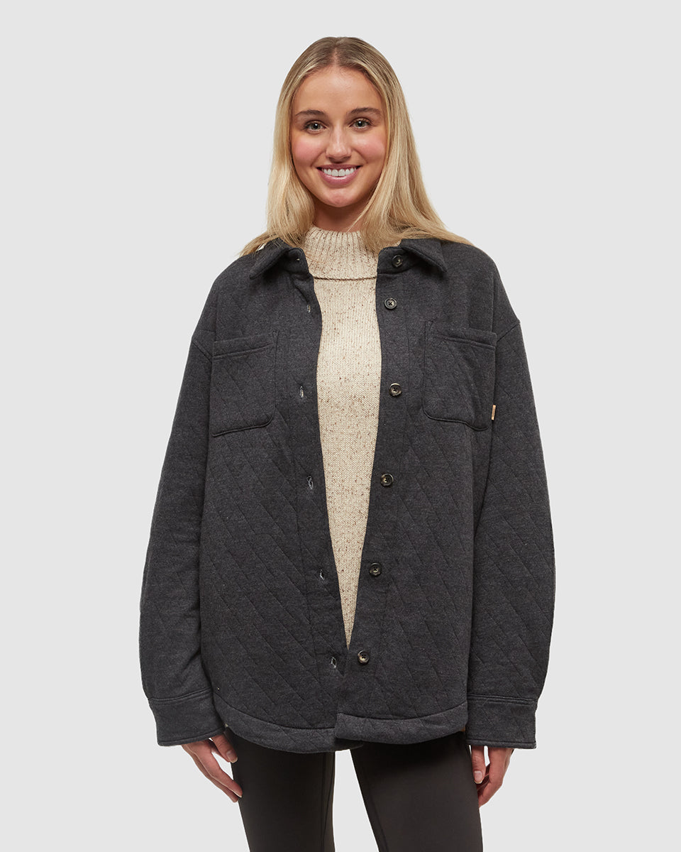 A woman with blonde hair smiles at the camera, wearing a dark grey quilted jacket over a cream-colored turtleneck sweater. The jacket is unbuttoned, revealing the sweater underneath. The background is a plain, light grey. The text overlay reads: "Warm when it’s cold. Water-repellent when it’s wet. Looks good dressed up or down." The ad creative focuses on the versatility and functional benefits of the jacket.