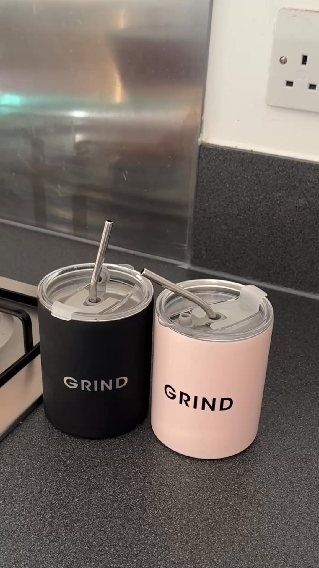 Two insulated tumblers, one black and one pink, sit on a kitchen counter. Both tumblers have the word "GRIND" printed on them in white text. Each tumbler has a metal straw inserted into its lid. The background shows a stainless steel backsplash and a white electrical outlet. The ad copy mentions a free gift with a subscription to coffee deliveries, using specific codes for the pink or black cup.