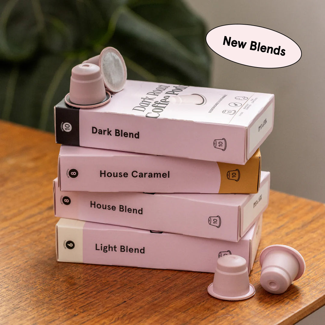 A stack of four pink boxes of coffee pods sits on a wooden table. The top box is labeled "Dark Blend," followed by "House Caramel," "House Blend," and "Light Blend." Two pink coffee pods are placed on top of the stack, and two more are on the table in front of the stack. An oval speech bubble in the top right corner contains the text "New Blends." The background shows a blurred green plant.