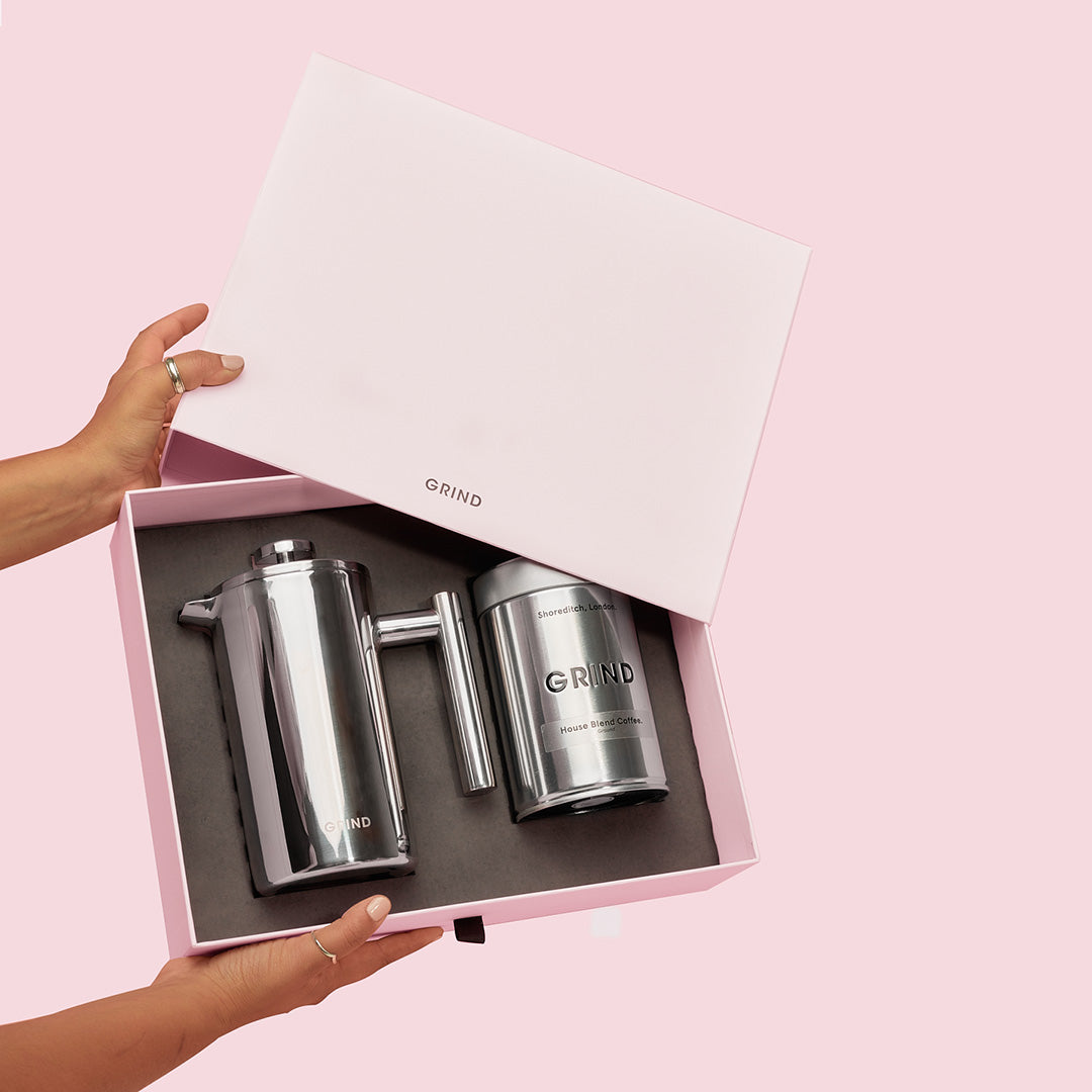 A light pink box is opened to reveal a shiny silver French press and a silver tin of coffee. The box and the products inside have the brand name "GRIND" printed on them. The lid of the box has "GRIND" printed on it in black text. The coffee tin has "Shoreditch, London" and "GRIND House Blend Coffee" printed on it. The background is a solid light pink color. The hook text "Your 50% OFF is waiting 👀" is overlaid on the image.