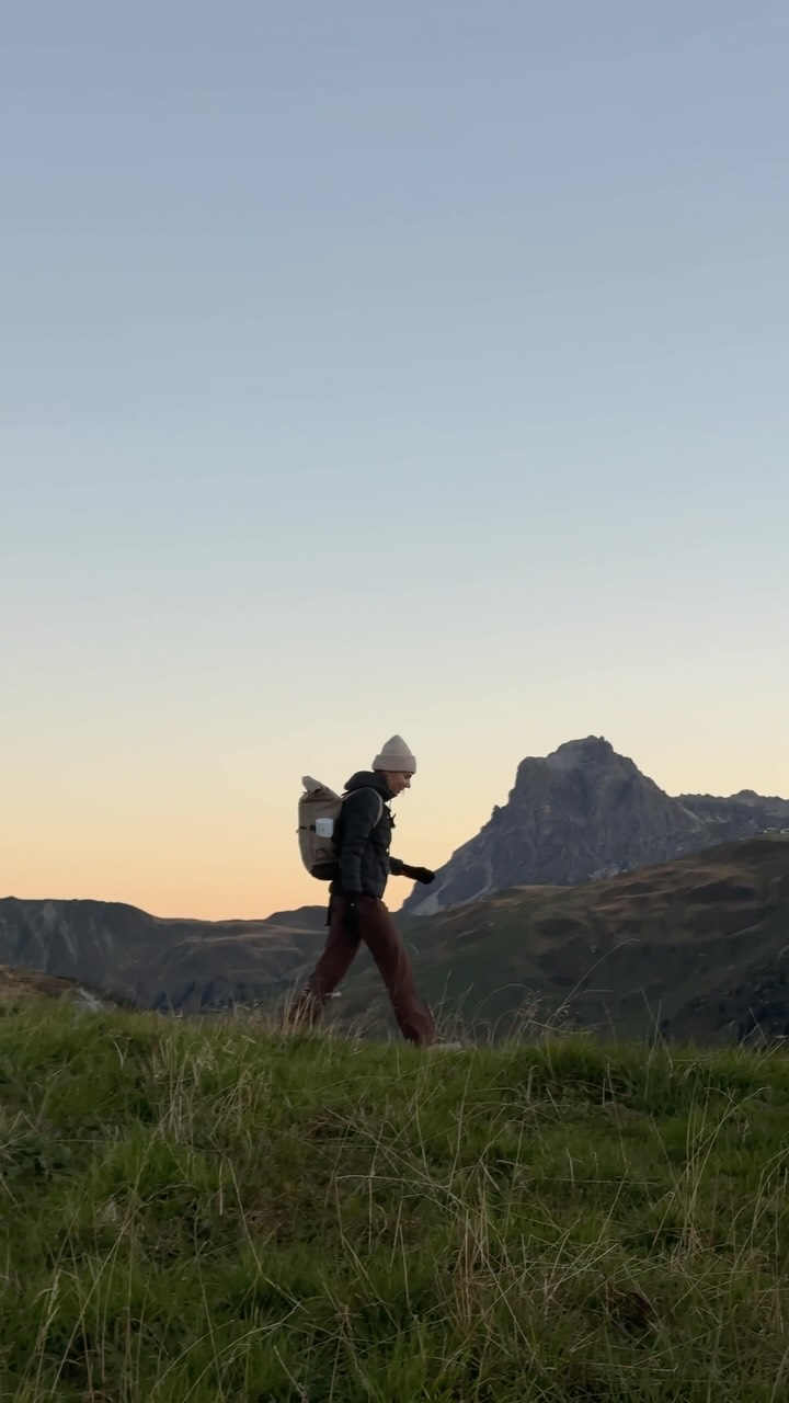 A person is walking across a grassy hill in the mountains at sunrise. They are wearing a beanie, jacket, and pants, and carrying a backpack. The sky is a gradient of pale blue and orange. The text overlay reads: "A crispy morning, self brewed coffee and watching the sun come up AD @mammut". The ad highlights the functionality of the LOOPINSULATION jacket in cold mountain conditions.