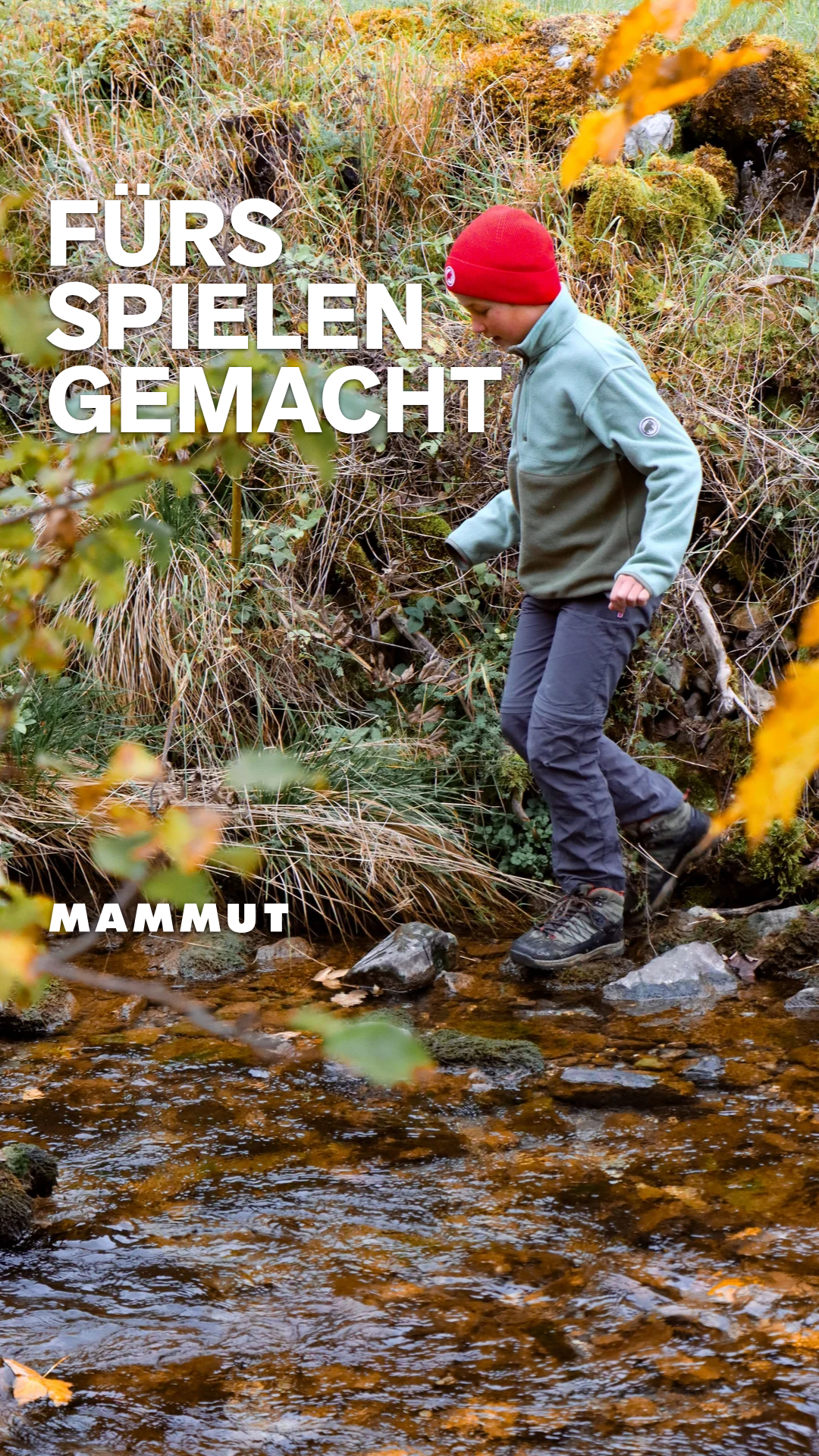 A young person wearing a red beanie and a light green fleece jacket walks across a shallow stream in a wooded area. Large white text overlays the image, reading "FÜRS SPIELEN GEMACHT" on the top left and "MAMMUT" on the bottom left. The scene is autumnal, with dry grass and yellow leaves visible. The text suggests the clothing is made for play and adventure.
