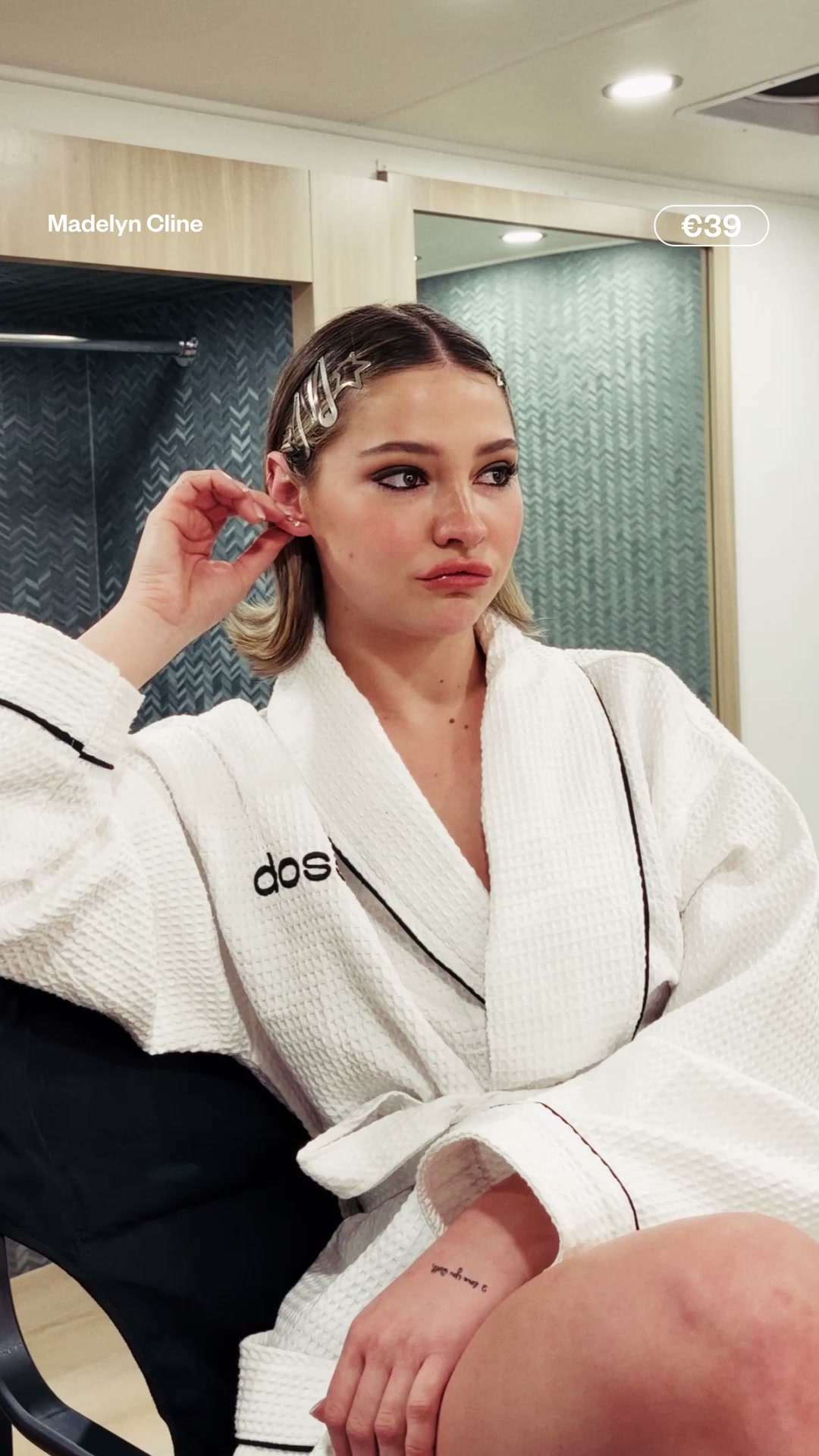Madelyn Cline is shown wearing a white bathrobe and adjusting her earring. Text overlays her image, stating "Madelyn Cline" in the top left corner and "€39" in a rounded rectangle in the top right. The background is a blurred interior with a tiled wall and a mirror. The overall visual suggests a behind-the-scenes or personal moment, with the celebrity endorsing a product.