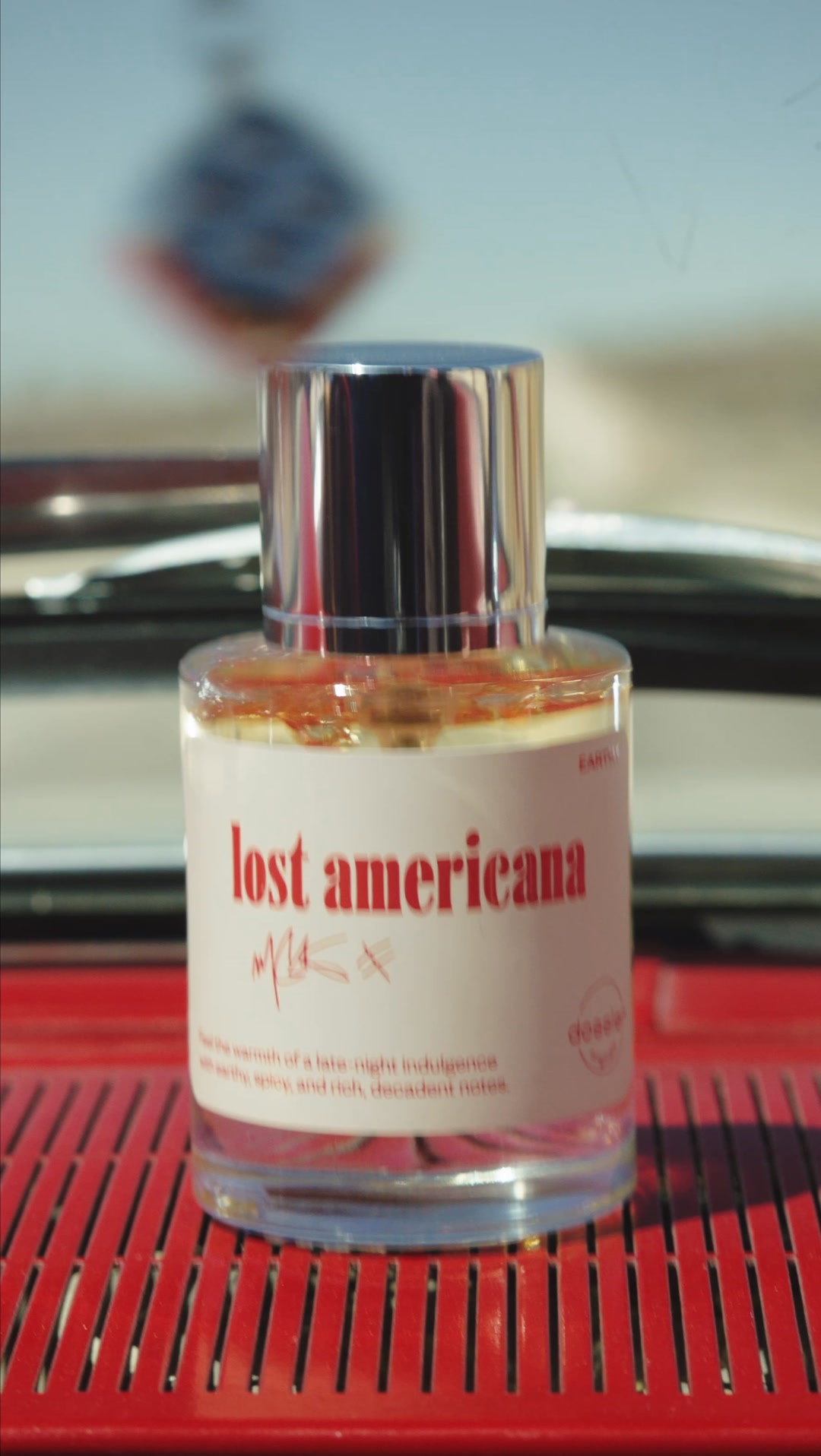 A close-up shot of a Dossier perfume bottle labeled "lost americana" is presented. The bottle contains a warm amber liquid and has a silver cap. The label also features handwritten-style text and a description of the scent. The bottle is placed on a red, slatted surface, with a blurred background suggesting an outdoor or car interior setting. The text on the bottle describes the fragrance as having earthy, spicy, and rich, decadent notes, evoking a sense of late-night indulgence.