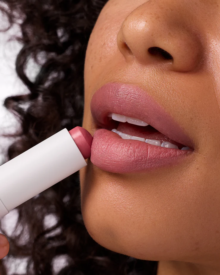 A close-up shot focuses on a person's lips as they apply a tinted lip balm. The balm tube is white with a pink tip, and the product itself is a soft pink shade. The person's lips are slightly parted, revealing white teeth, and their skin tone is warm. The background is blurred, with hints of dark curly hair visible on the left side. The on-screen text reads: "Made for sunny plans." The copy below the image explains that the tinted lip balm offers hydration, color, and SPF 30 for use throughout the day.