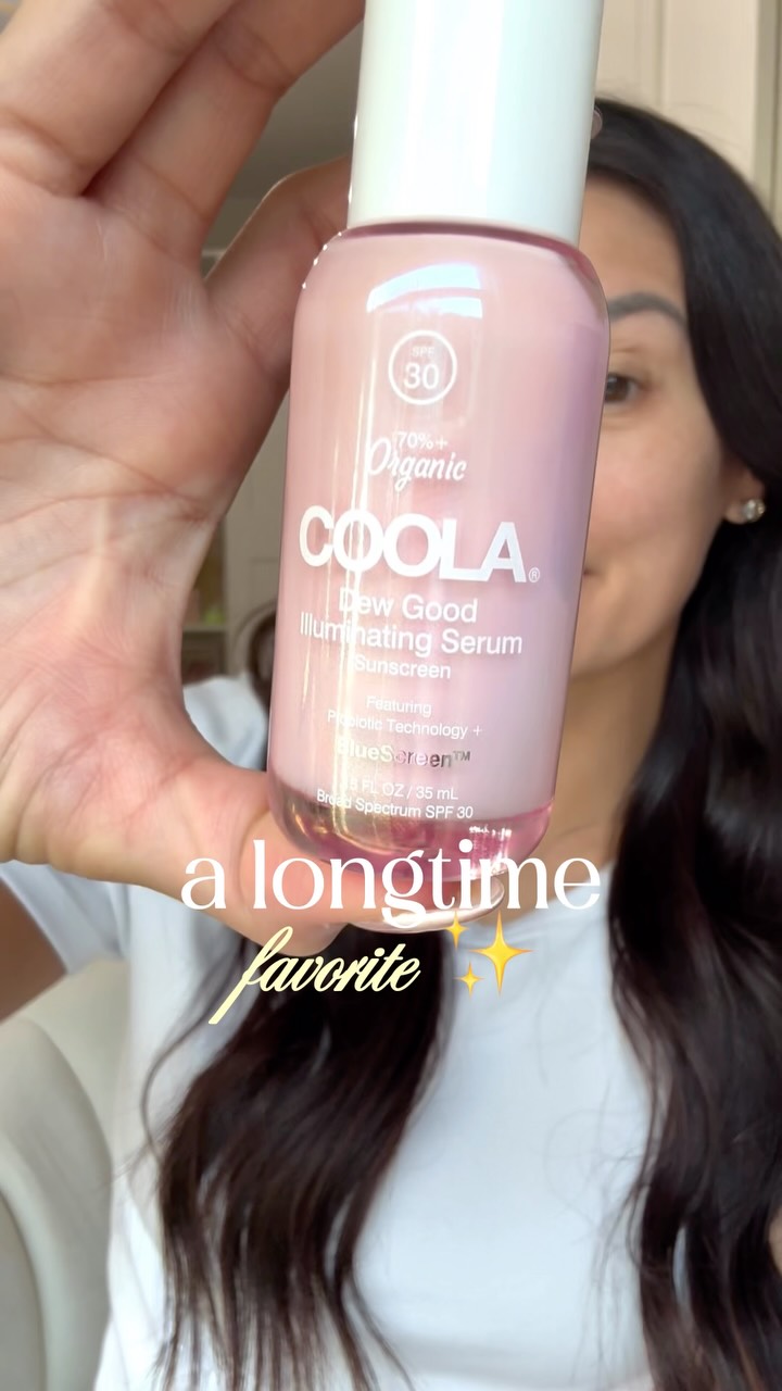 A person holds up a bottle of COOLA Dew Good Illuminating Serum Sunscreen SPF 30. The bottle is pink and has white text. The person is smiling slightly and looking at the camera. Overlay text in a cursive font reads "a longtime favorite" with sparkles. The background is blurred and appears to be indoors. The creative highlights the product's ability to provide a glowy look with minimal effort and its SPF protection.