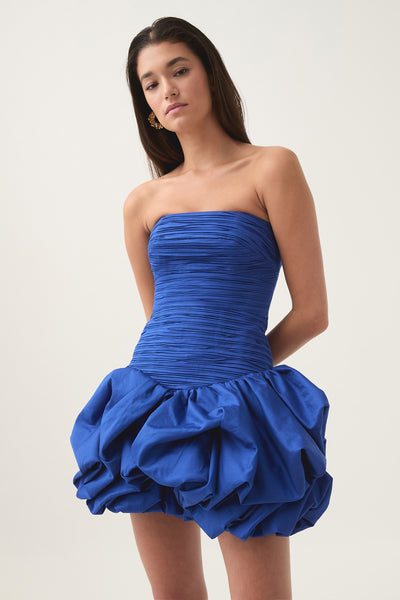 A model is shown from the waist up, wearing a vibrant blue strapless dress with a ruched bodice and a voluminous bubble skirt. She has long, dark hair and is looking directly at the camera with a neutral expression. Gold earrings are visible. The background is a plain, light-colored studio setting. Text overlayed on the image reads: "Caught your eye? Make it yours. Find it at Aje."