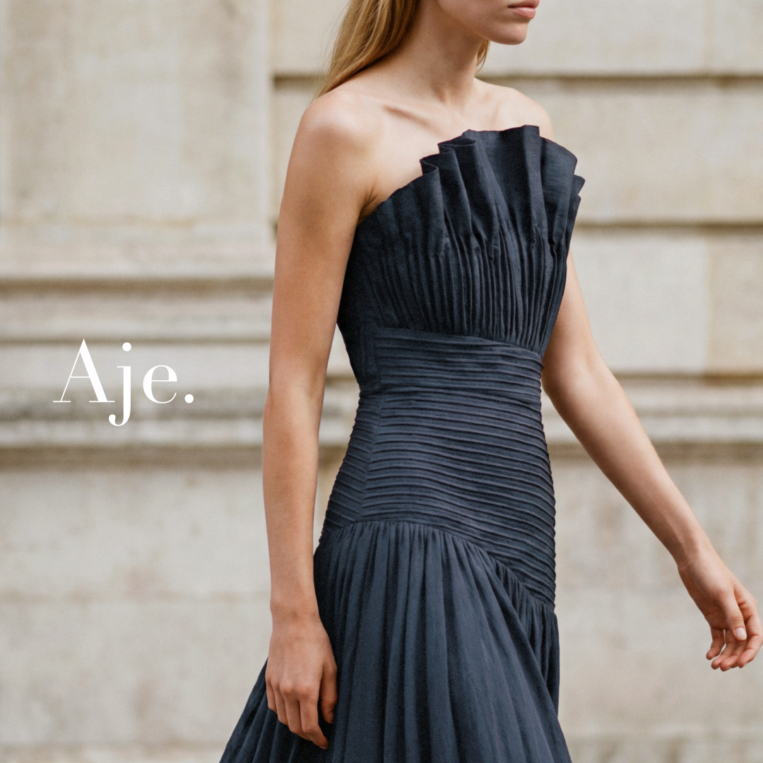 A close-up, side-profile shot of a woman wearing a dark, strapless, ruched gown. The text "Aje." is overlaid in a large, white, serif font on the left side of the image. The background is a blurred, light-colored stone building. The woman is looking to the right and her right arm is extended slightly, with her hand open. The image focuses on the texture and design of the dress.