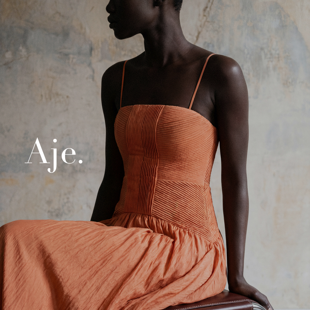A woman of color is seated, wearing an orange, textured, strappy dress. The text "Aje." is overlaid on the left side of the image. The background is a textured, neutral-colored wall. The overall aesthetic is sophisticated and highlights the intricate details of the garment, suggesting a moment of refined arrival and feminine elegance.