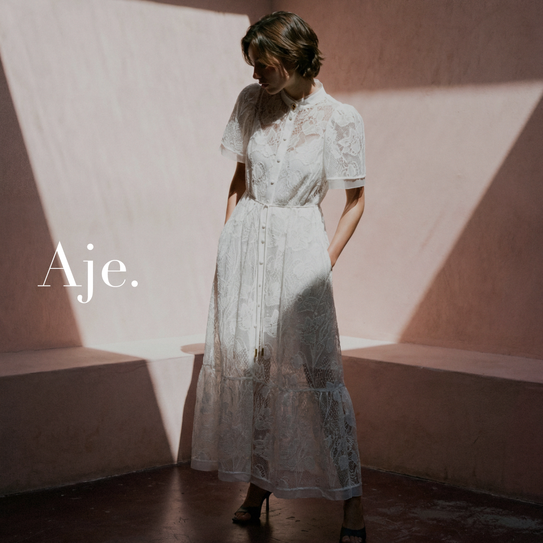 A woman stands in a room with pink walls and dramatic shadows cast by sunlight. She is wearing a white lace midi dress with short sleeves and a belted waist. The brand name "Aje." is written in large, elegant white font on the left side of the image. The text overlay introduces the Pippa Lace Midi dress as a versatile piece for day to night styling.