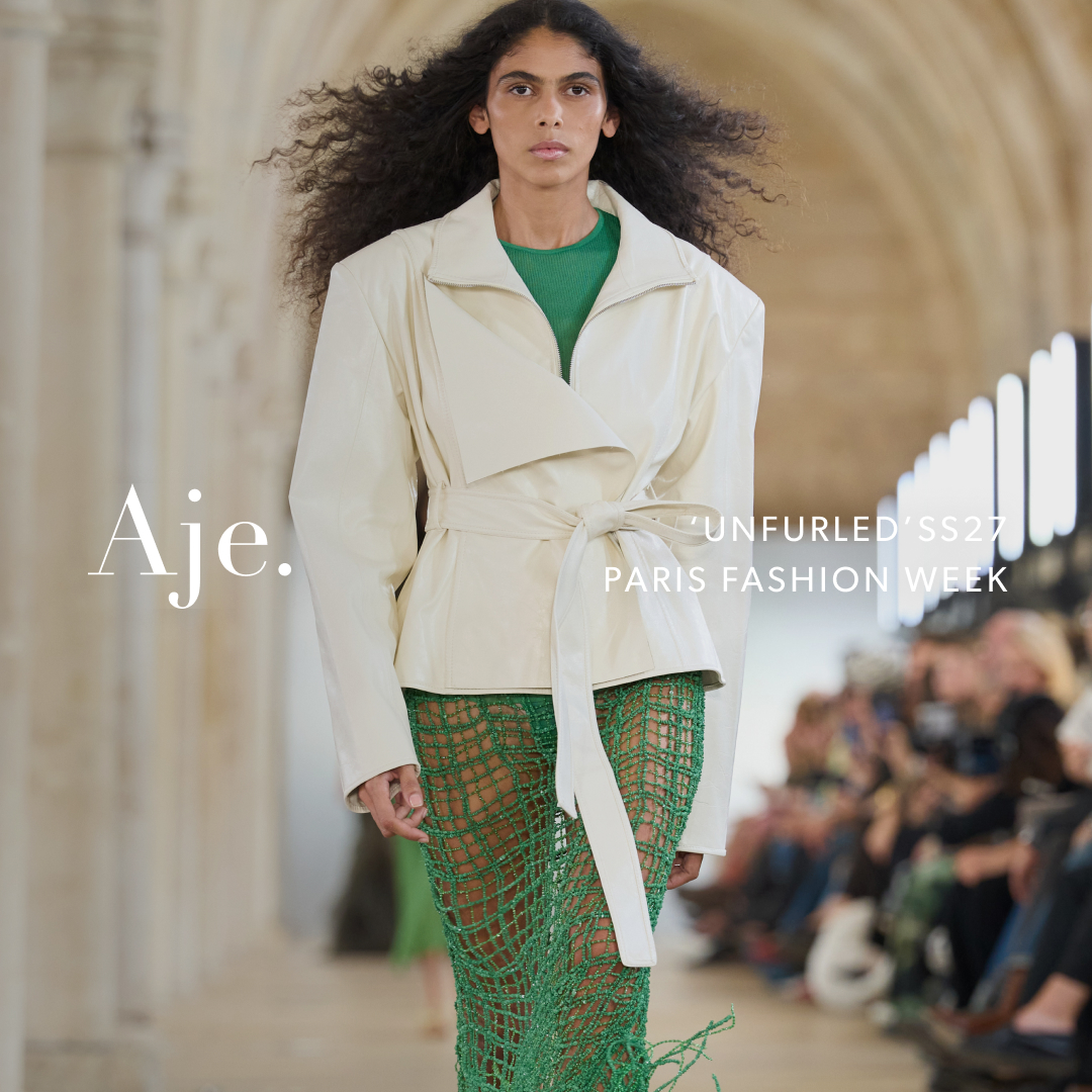 A female model with long, dark, curly hair walks down a runway. She is wearing a cream-colored, belted jacket over a green top and a green, netted skirt. White text on the left side of the image reads "Aje." White text on the right side of the image reads "'UNFURLED' SS27 PARIS FASHION WEEK." The background shows an indoor runway with blurred audience members.