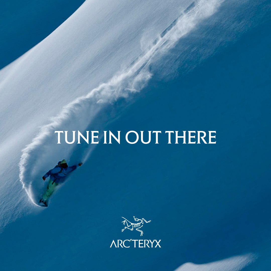 A snowboarder carves down a steep, snow-covered mountain, kicking up a spray of powder. White text overlaid on the image reads "TUNE IN OUT THERE." Below the text, the Arc'teryx logo is visible. The overall scene evokes a sense of adventure and connection with nature, suggesting the brand's gear is suited for such experiences.