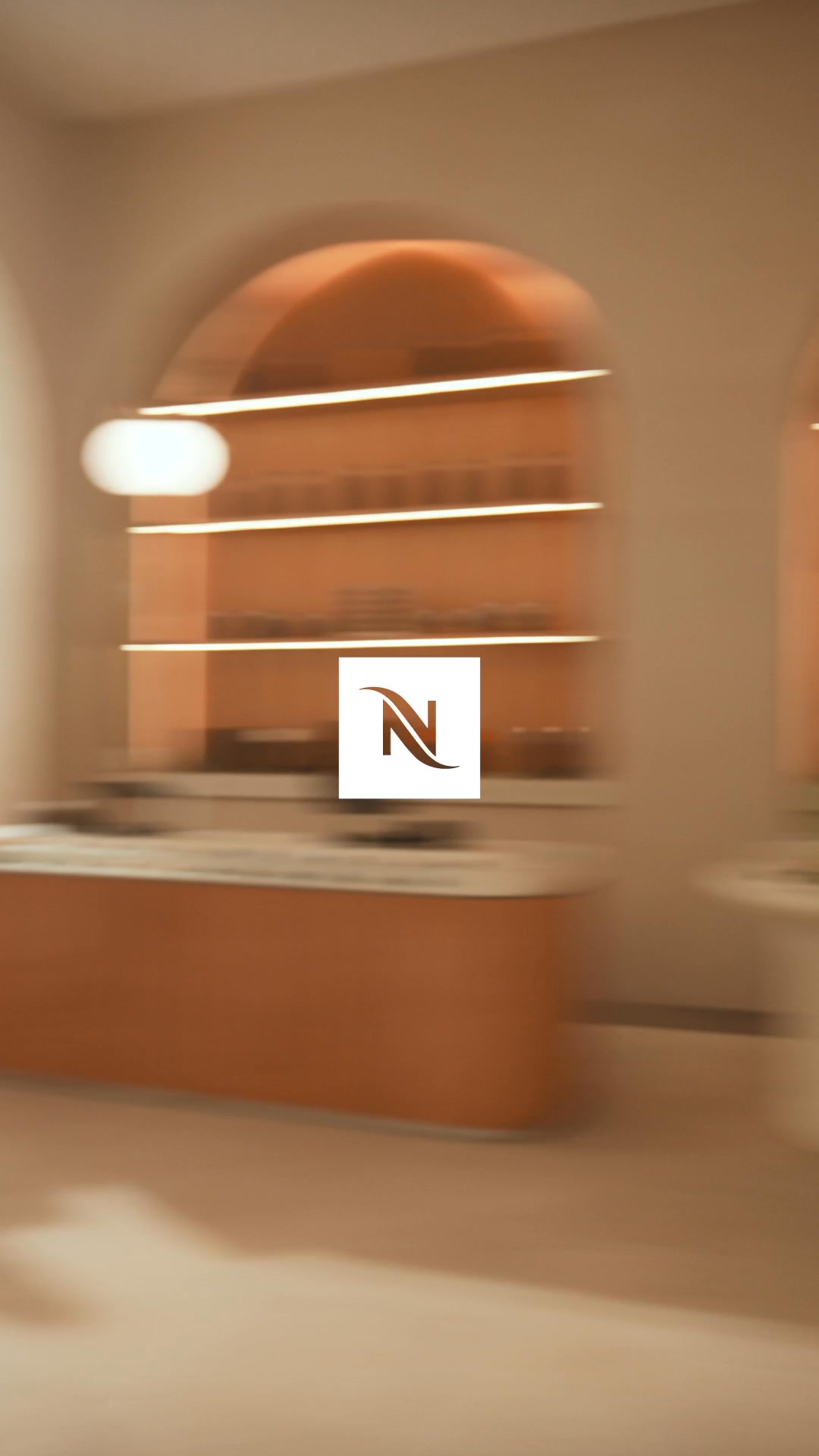 The creative shows a blurred, interior shot of a modern Nespresso store. The focus is on a curved counter and shelves stocked with coffee products, illuminated by warm lighting. A Nespresso logo is prominently displayed in the center of the frame. The text overlay invites the viewer to follow Dua Lipa enjoying a Nespresso iced latte, highlighting its cool and refreshing taste.