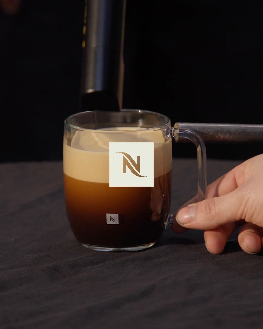 A close-up shot shows a clear glass mug filled with dark coffee and a thick layer of creamy foam on top. The Nespresso logo is prominently displayed in a light beige square on the foam, and a smaller Nespresso logo is visible on the coffee itself. A hand is seen holding the mug's handle, with a coffee machine's spout visible above the mug, suggesting the coffee is being freshly brewed. The background is dark and out of focus, drawing all attention to the coffee and the brand.