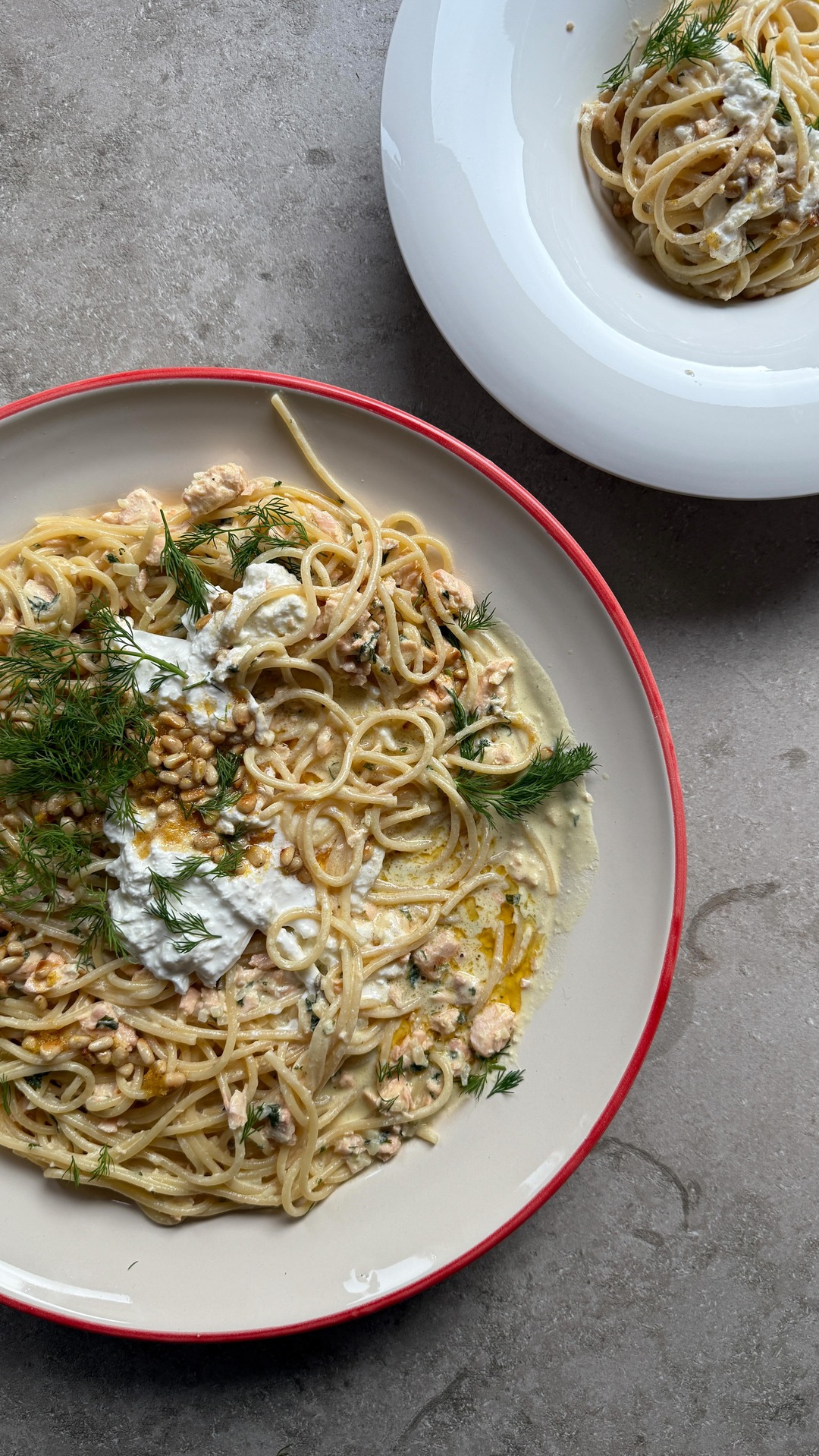 A top-down shot of two plates of spaghetti with salmon and burrata. The main plate in the foreground is a close-up of the pasta, which is topped with dollops of creamy burrata, chopped salmon, pine nuts, and fresh dill. The sauce appears to be a creamy, light-colored sauce. A second white plate with pasta is visible in the background. The text overlay is the hook, which is a personal endorsement of the dish and its texture.