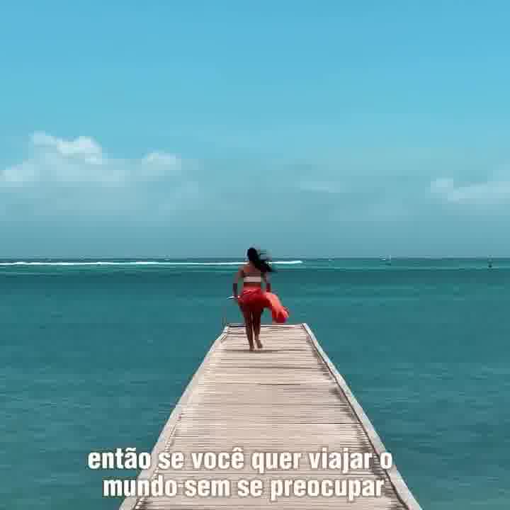 A person walks away from the camera down a wooden pier towards a vast blue ocean under a clear sky. The text on the screen reads, "so if you want to travel the world without worrying". The image evokes a sense of freedom and wanderlust, suggesting a carefree travel experience.
