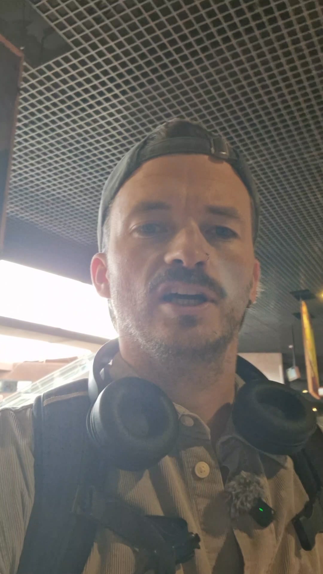 A man wearing a baseball cap and headphones is filming himself in a selfie-style video. He has a beard and a nose ring. He is wearing a collared shirt and a backpack strap is visible. The background appears to be an indoor public space with a grid-like ceiling. Text overlays the video, stating "Find eSIMs for 200+ countries and regions" and "✈️ Perfect for holiday travel 🛜 Stay connected with Airalo!"