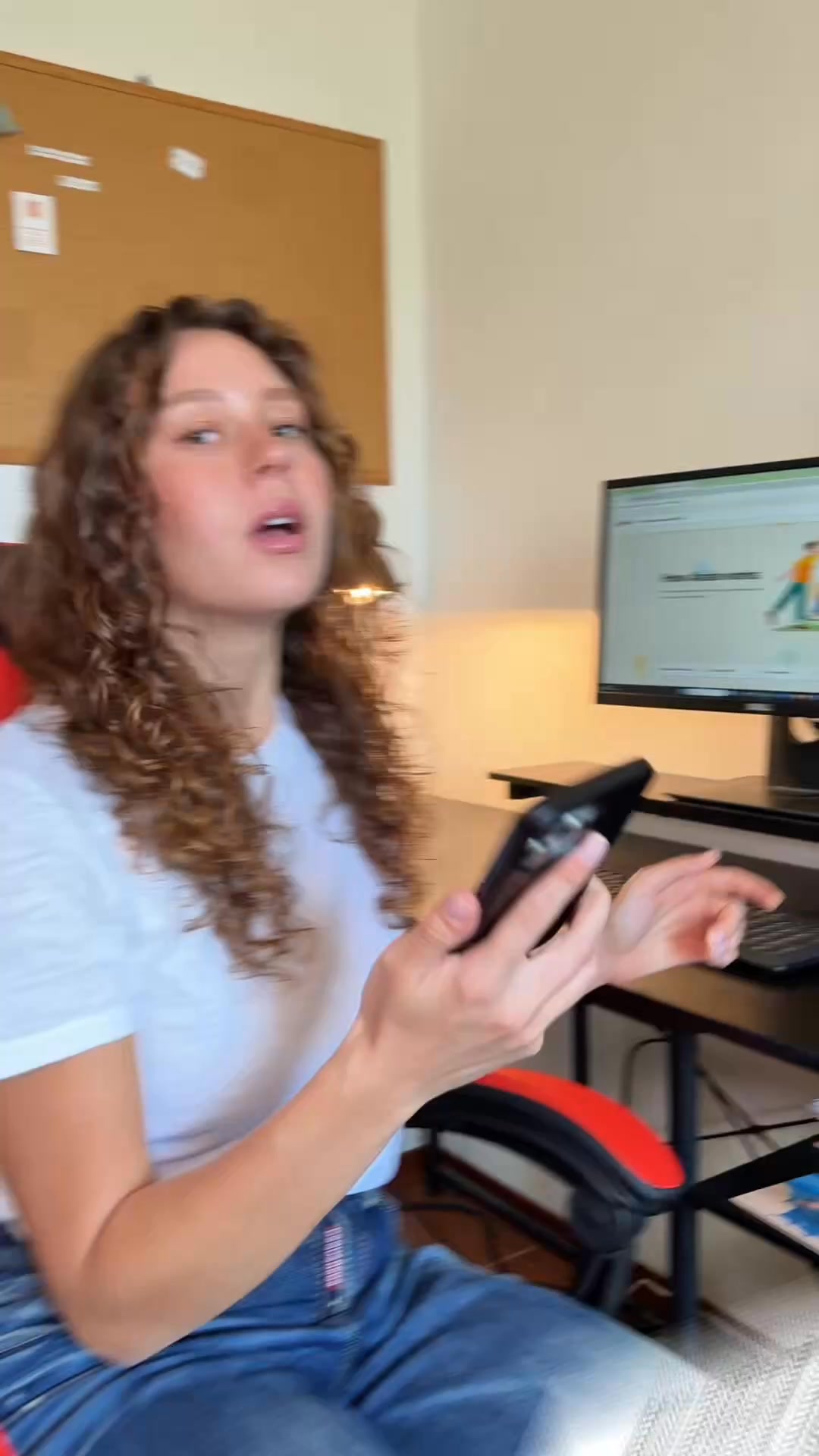 A young woman with curly brown hair, wearing a white t-shirt and blue jeans, is sitting in an office chair and holding a smartphone. She is looking towards the camera with an expressive look on her face. Behind her, a computer monitor displays a webpage with graphics of people. The overall scene suggests a casual work or study environment. The text overlay encourages exploration and sharing experiences.